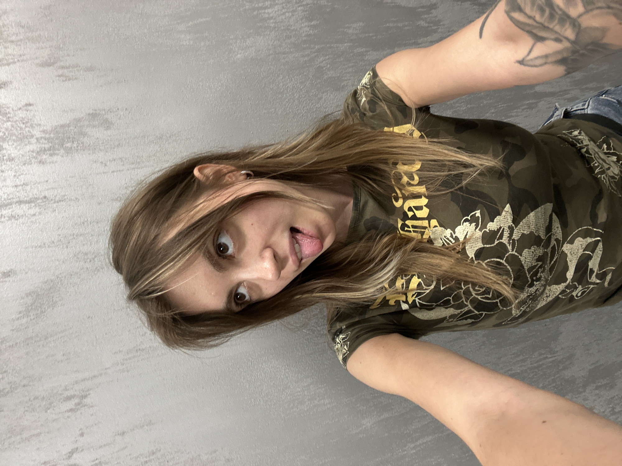 The image shows a young woman with long, brown hair and light brown eyes. She is sticking her tongue out at the camera and has a playful expression. She is wearing a camouflage t-shirt with gold lettering and a flower design. There is a tattoo visible on her arm. She appears to be indoors, in front of a textured grey wall. The lighting suggests it is daytime. The orientation of the image is rotated 90 degrees counter-clockwise. The text on her shirt is partially obscured but appears to be a word or phrase in a stylized font. Based on the location context provided, this image was taken in Moscow, Russia.