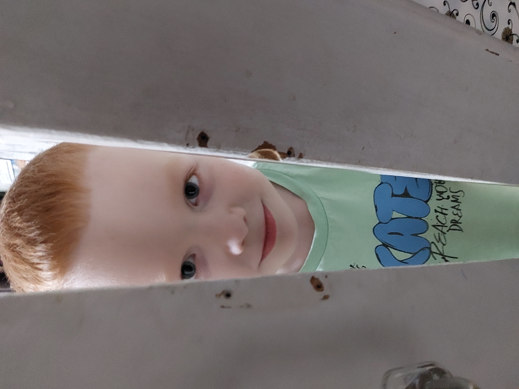 A red-haired child wearing a light green t-shirt with blue lettering is peeking through a gap in what appears to be a painted white wooden structure, potentially a fence or railing. The setting is ambiguous, no location in Chaikovsky, Russia is indicated. The child's face is close to the camera, with their eyes looking directly at the viewer. The lighting is soft and diffused. The background is partially visible through the gap, showing a wall or surface with a decorative pattern.