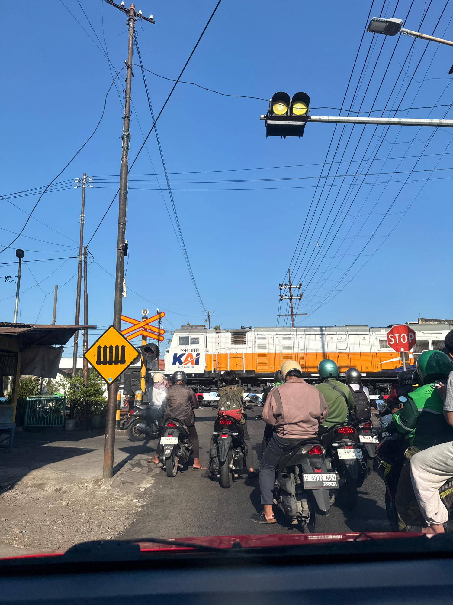 The image depicts a street scene in Surabaya, Indonesia, near a railway crossing. Several motorcycles are stopped in traffic, waiting for a train. The train, white with orange accents, is a KAI (Kereta Api Indonesia) locomotive. A yellow railroad crossing sign with a depiction of train tracks is visible on the left. Traffic lights showing yellow are also present, indicating an approaching train or an active crossing. The sky is clear and blue, with numerous overhead power lines crisscrossing the frame. People on the motorcycles are wearing helmets and casual clothing. The environment suggests an urban setting with a mix of infrastructure and everyday life.
