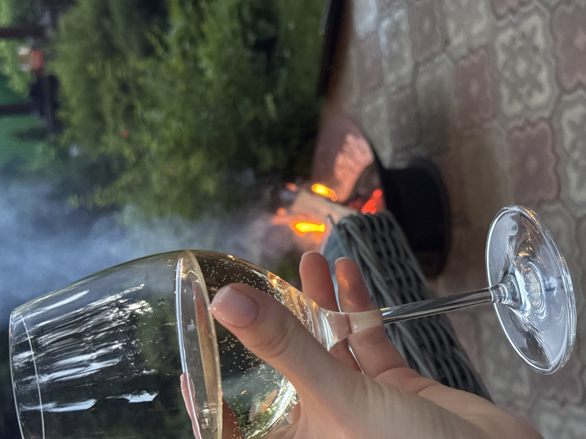 A person's hand holds a glass of white wine in front of a backyard fire pit. The hand, with manicured nails, gently grips the stem of the wine glass. The glass contains a pale yellow liquid with numerous small bubbles rising to the surface. In the background, a fire burns in a dark metal fire pit, with flames illuminating pieces of wood and smoke rising into the air. A woven basket is positioned near the fire pit. The ground is paved with interlocking tiles. Lush green foliage forms the backdrop, suggesting an outdoor setting in a residential area. The image appears to be taken at dusk, as indicated by the soft lighting and the presence of the fire. The specific location of Tomilino, Russia, is not discernible from the visual elements in the image itself.