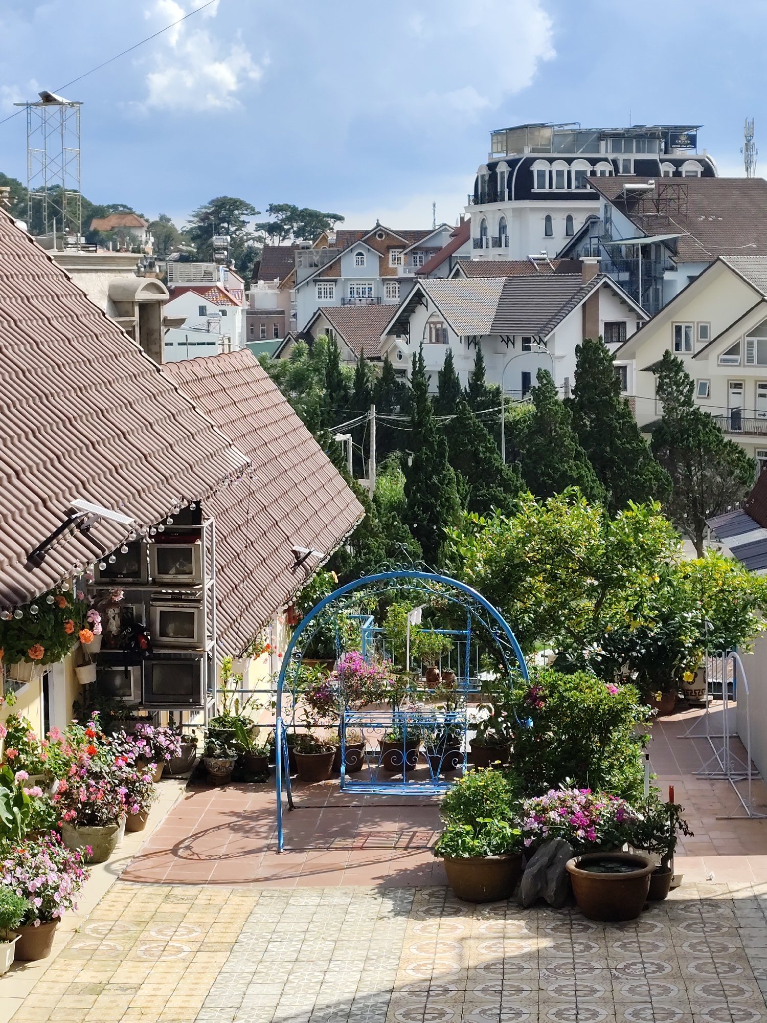 This is a medium-shot, eye-level photograph taken outdoors, likely on a sunny day with some clouds. The scene is a charming courtyard or balcony filled with potted plants and flowers, overlooking a hillside town with numerous houses and buildings. The architecture of the town is varied, featuring a mix of tiled roofs, modern structures, and some with ornate window details. In the foreground, a tiled patio area is visible, with a blue metal swing set providing a focal point. The plants are lush and in bloom, with vibrant pink and orange flowers adorning the space, creating a cheerful and inviting atmosphere.
To the left, a portion of a house with a red-tiled roof dominates the frame, with a shelf unit holding several old televisions against its wall. Some decorative string lights are visible along the edge of the roof. The overall impression is one of a peaceful, well-tended outdoor space in a hilly, picturesque town. The lighting suggests it is daytime, perhaps late morning or early afternoon, given the clear sky and shadows. No people are visible in the frame, and the focus is on the interplay of architecture, nature, and decorative elements. A small sign with Vietnamese text is visible on the rooftop of a prominent building in the background.