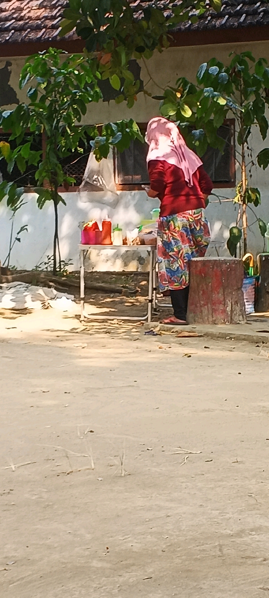 A woman wearing a pink hijab and a red sweater is standing behind a small table. She is wearing a colorful, floral patterned skirt and black pants. On the table are various items, including a pink basket, two squeeze bottles, and other containers with food or condiments. The scene appears to be outdoors, possibly in front of a building with a tiled roof and white walls. There are plants and trees in the background. The ground in front of the table is dirt or sand. This scene is located in the city of Penanggungan, Indonesia.