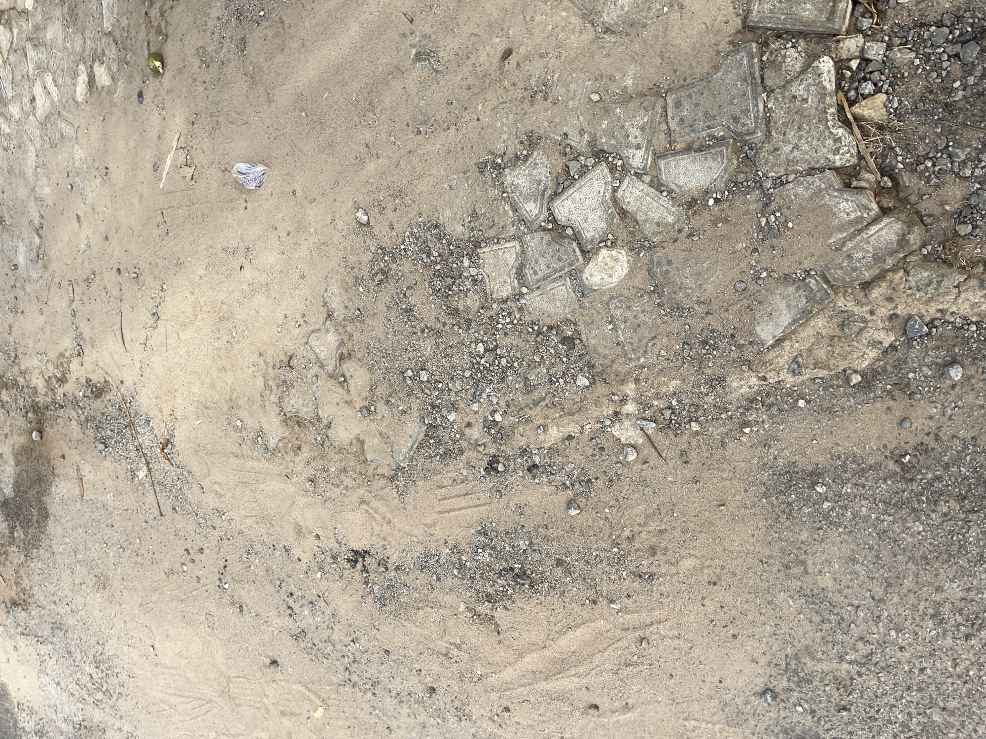 The scene is a close-up of a ground surface, likely a section of an unpaved road or construction site. The surface is mostly composed of sand and gravel. Disrupted pieces of concrete pavement or tiles are scattered across the scene, some broken into smaller fragments. A small piece of light blue or white litter lies on the sandy area. There are patterns in the sand, possibly from the passage of a tire or movement of wind.
It's impossible to confirm the specific location in Abuja, Nigeria without further information.