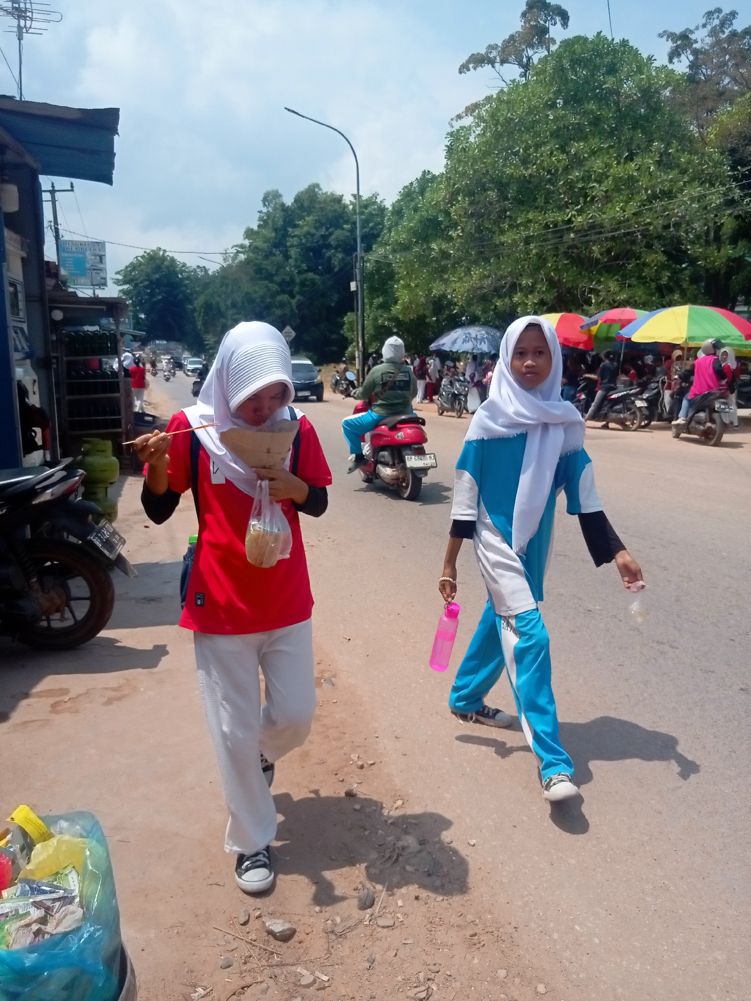Two young females are walking on a street in Batam, Indonesia, on a sunny day. The female on the left is wearing a red shirt, white pants, and a white hijab. She is holding a paper cone of food and chopsticks, appearing to eat. The female on the right is wearing a blue and white tracksuit and a white hijab. She is holding a pink water bottle and a plastic bag. They are walking towards the right side of the frame, on a road with traffic, including motorcycles and cars. In the background, there are trees, buildings, and colorful umbrellas. A blue trash bag filled with items is visible on the left side of the image, near a parked motorcycle.