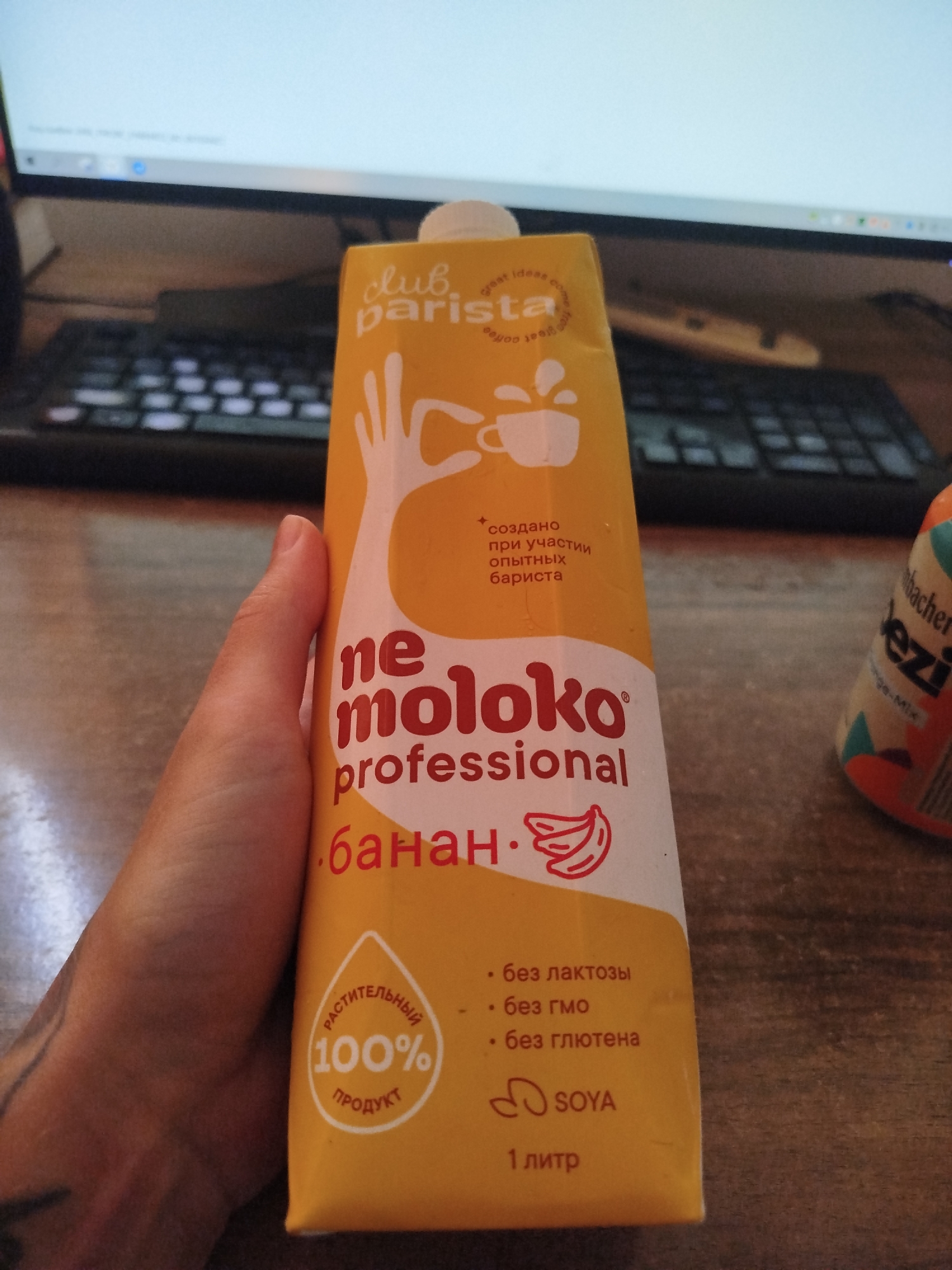 A person's hand is holding a bright yellow carton of "Ne Moloko Professional" banana-flavored plant-based milk. The carton has red lettering and graphics, including a stylized drawing of a hand and a coffee cup, and Russian text indicating it is made with the participation of experienced baristas, is lactose-free, GMO-free, gluten-free, and is a soy product. The carton also states it is 100% plant-based product and contains 1 liter. In the background, a computer monitor and keyboard are visible, suggesting the photo was taken indoors, likely at a desk. A partial view of another beverage carton, possibly from "Dr. Kargen" with "ezzi" in large letters, is also visible to the right of the main carton. The overall lighting is neutral.