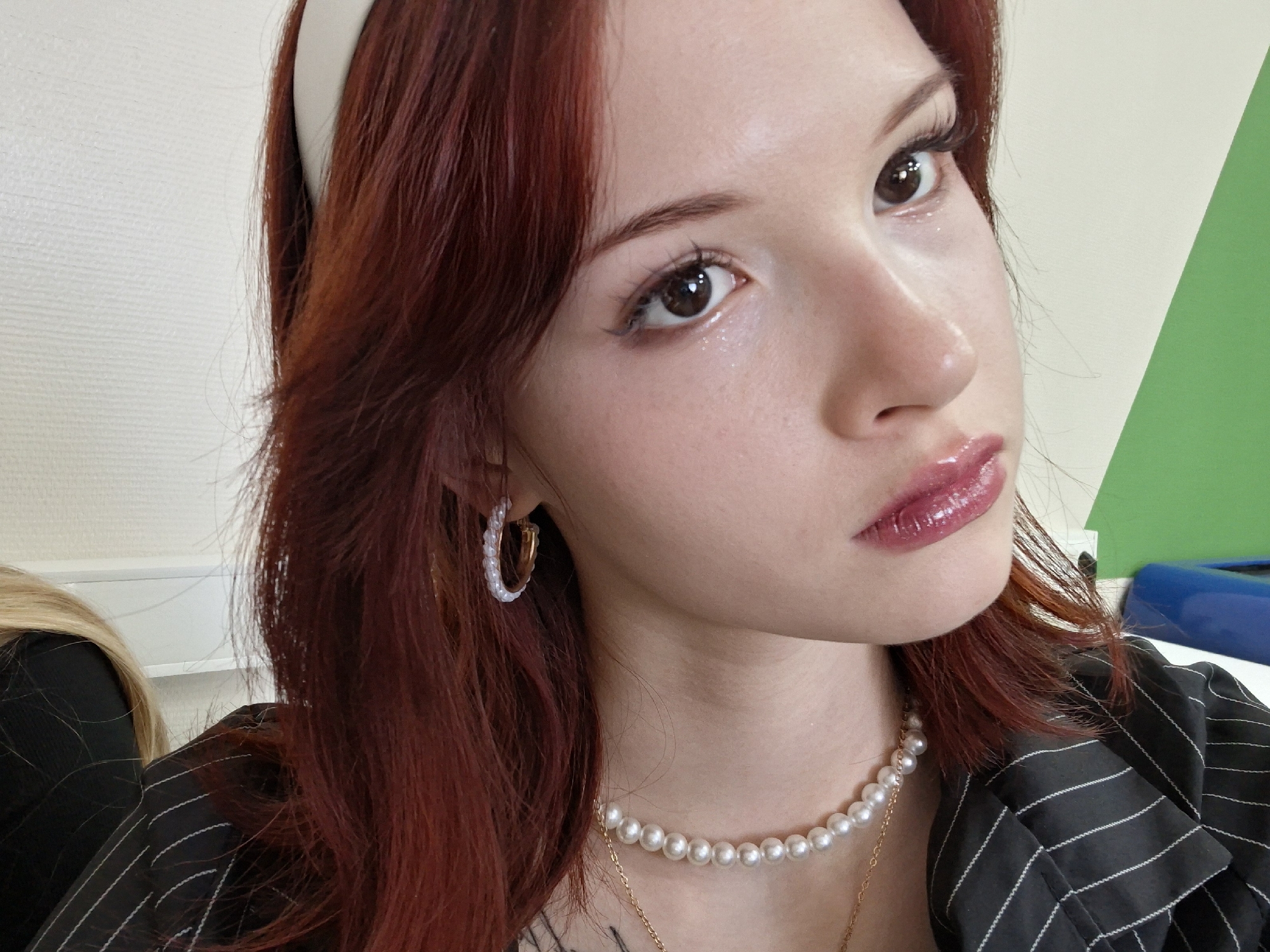 A close-up, high-angle shot depicts a woman with red hair, fair skin, and light makeup, including winged eyeliner and glossy lips. She is wearing a white headband, pearl earrings, and a pearl necklace. Her clothing appears to be a black pinstriped blazer. A portion of another person with blonde hair is visible in the lower left corner. The background includes a white wall and a green and blue surface. There is no evidence to determine location in Chaikovsky, Russia.