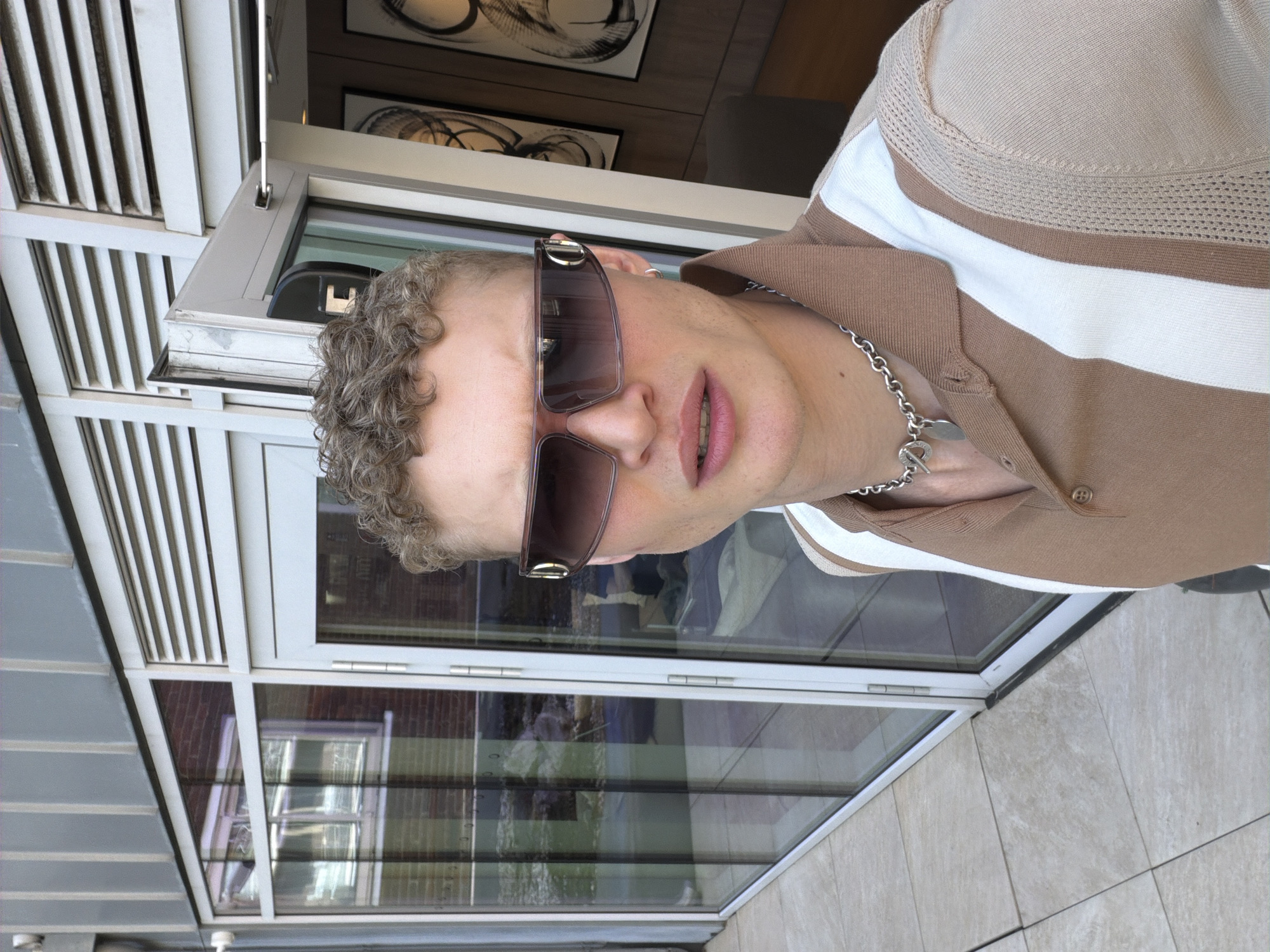 A man with fair skin, curly light-brown hair, and sunglasses is posing for a photo. He is wearing a tan and white striped shirt and a silver chain necklace. The background features a building with a gray exterior and large windows. Through the windows, a brick building and other architectural elements are visible. The presence of a white awning suggests the location is outdoors, possibly on a patio or deck.