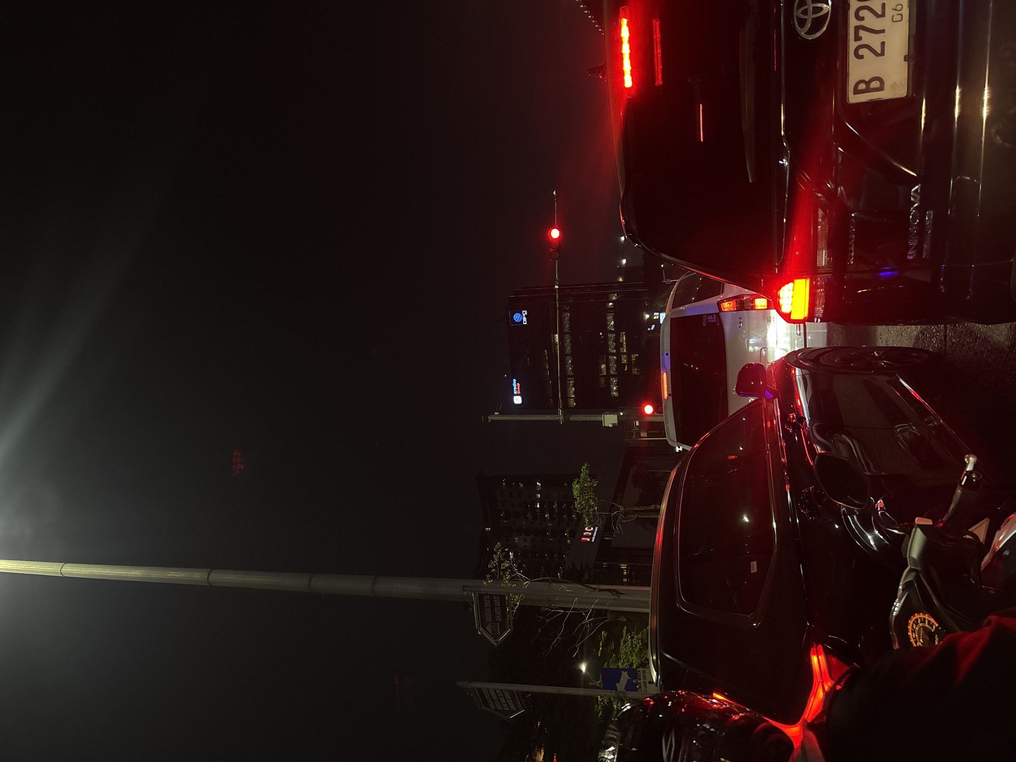 The image depicts a traffic jam at night in West Jakarta, Indonesia. Several cars are visible, including a black SUV in the foreground with its brake lights illuminated. A motorcycle is also partially visible on the left. In the background, tall buildings with lit windows are present, suggesting an urban environment. A traffic light displays a red signal. Street signs are visible on a lamppost, indicating directions to "MUSEUM TERBAIK JAKARTA BARAT". The overall scene is characterized by the darkness of night, illuminated by vehicle lights and building lights.