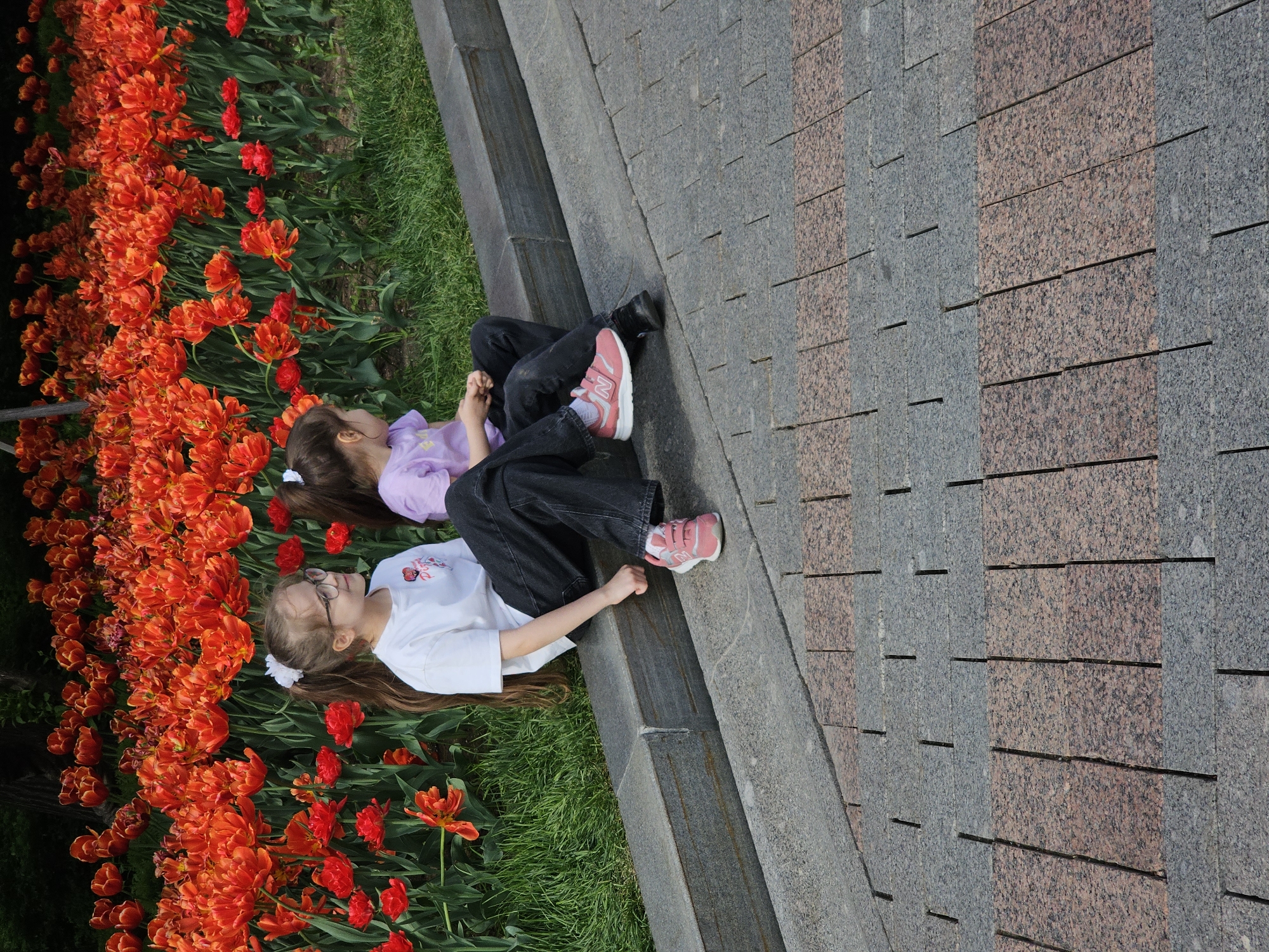 Two young girls are lying side-by-side next to a flower bed filled with red tulips, in what appears to be a city park. One girl wears a white shirt, black jeans, glasses, and pink sneakers. The other girl wears a purple shirt and black jeans and black shoes. They are positioned between the flower bed and a paved area that looks like a walkway. The pavement has gray and red bricks. The city is unknown, but the scene suggests a public space with landscaping.