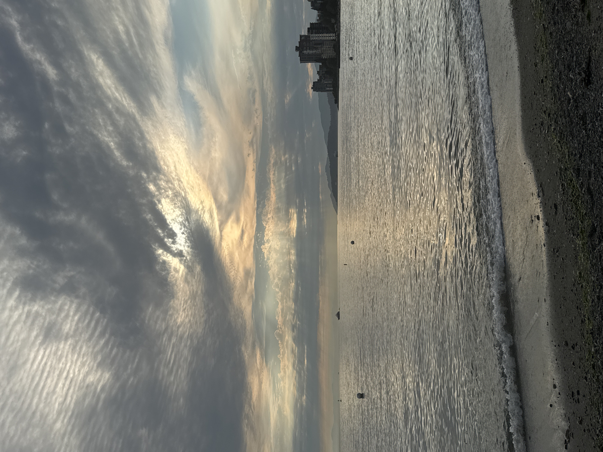 A wide shot captures a serene seascape at dusk. The sky is a canvas of dramatic clouds, with streaks of pale orange and yellow light breaking through, casting ethereal rays onto the water. The sea itself is calm, with gentle waves lapping at a pebbly shore. In the distance, a city skyline is visible, featuring several tall buildings that contrast with the softer, mountainous backdrop. A few small buoys dot the water, and a boat can be seen sailing towards the horizon. The overall mood is tranquil and atmospheric, hinting at a coastal urban environment.