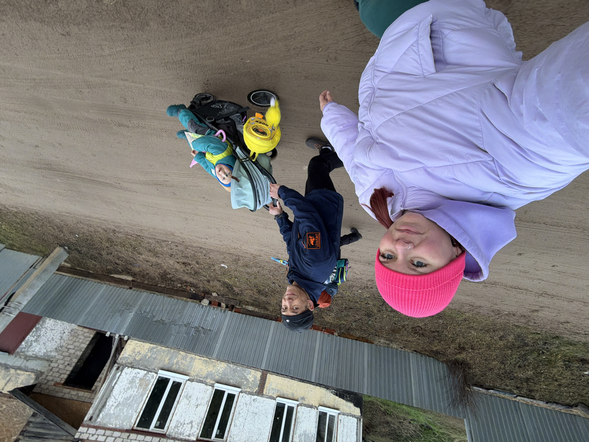 A woman in a pink beanie and light purple puffy jacket stands next to a man in a dark blue jacket, both looking at the camera. The man is pushing a stroller with a child bundled in blue in it. A yellow bag resembling a duck is attached to the stroller. They appear to be on a dirt road next to a dilapidated building with barred windows. The scene is located in what could be Chaikovsky, Russia.