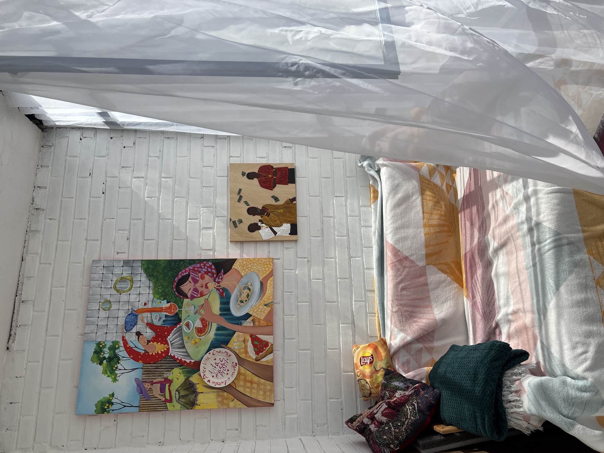 The scene depicts the interior of a room with white brick walls. Two paintings are hanging on the wall: one depicting a group of people eating and drinking, and the other depicting two men and a pile of money. A bag of Lay's potato chips sits on the floor next to a floral pillow and a dark green scarf. The room is covered in white plastic sheeting.
The image does not contain information confirming the location in the city of Krasnoyarsk, Russia.