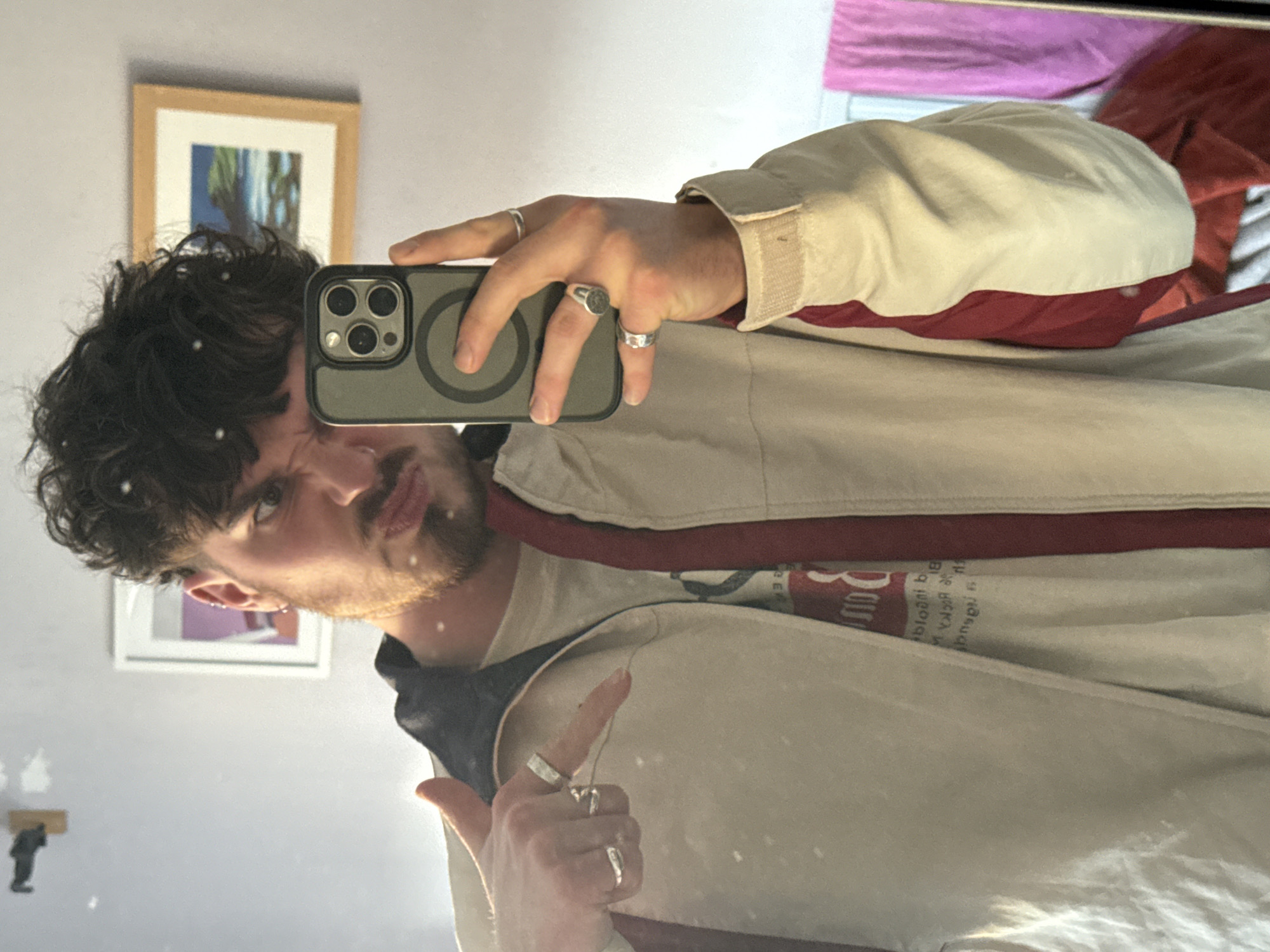 A male is taking a selfie in a mirror with a smartphone. He is wearing a beige jacket with red trim over a t-shirt. The man has curly dark hair and a beard. He has multiple rings on his fingers. Behind him, two framed pictures are hanging on a light-colored wall. In the upper right corner of the image, a pink and a red item are visible. The location is Unknown, Unknown.