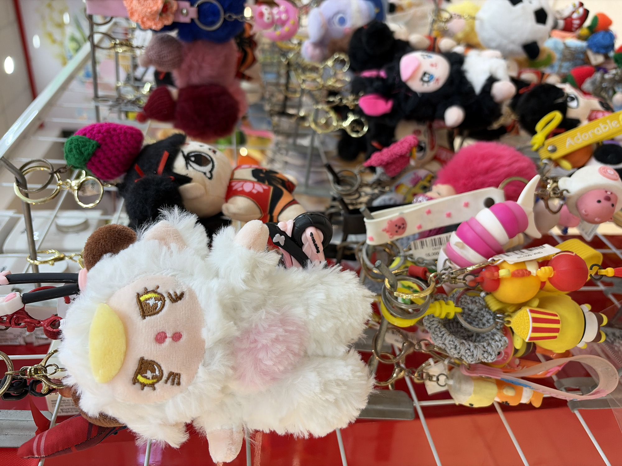 The image shows a close-up view of a display of keychains and plush toys in a store. The items are hanging on a metal grid, with many colorful and character-themed keychains. The most prominent item in the foreground is a fluffy white lamb plush toy with yellow eyes and a pink chest. Other keychains include a knitted strawberry, various cartoon characters, and animal-themed plushies like a black and white panda. The background is slightly blurred but suggests a retail environment with shelves and more merchandise. The scene is brightly lit, likely from overhead store lighting. The overall impression is a vibrant and whimsical collection of trinkets, typical of a gift shop or souvenir store. The items are all keychains, suggesting they are intended for decorative or functional use attached to keys or bags. There are no people visible in the image.