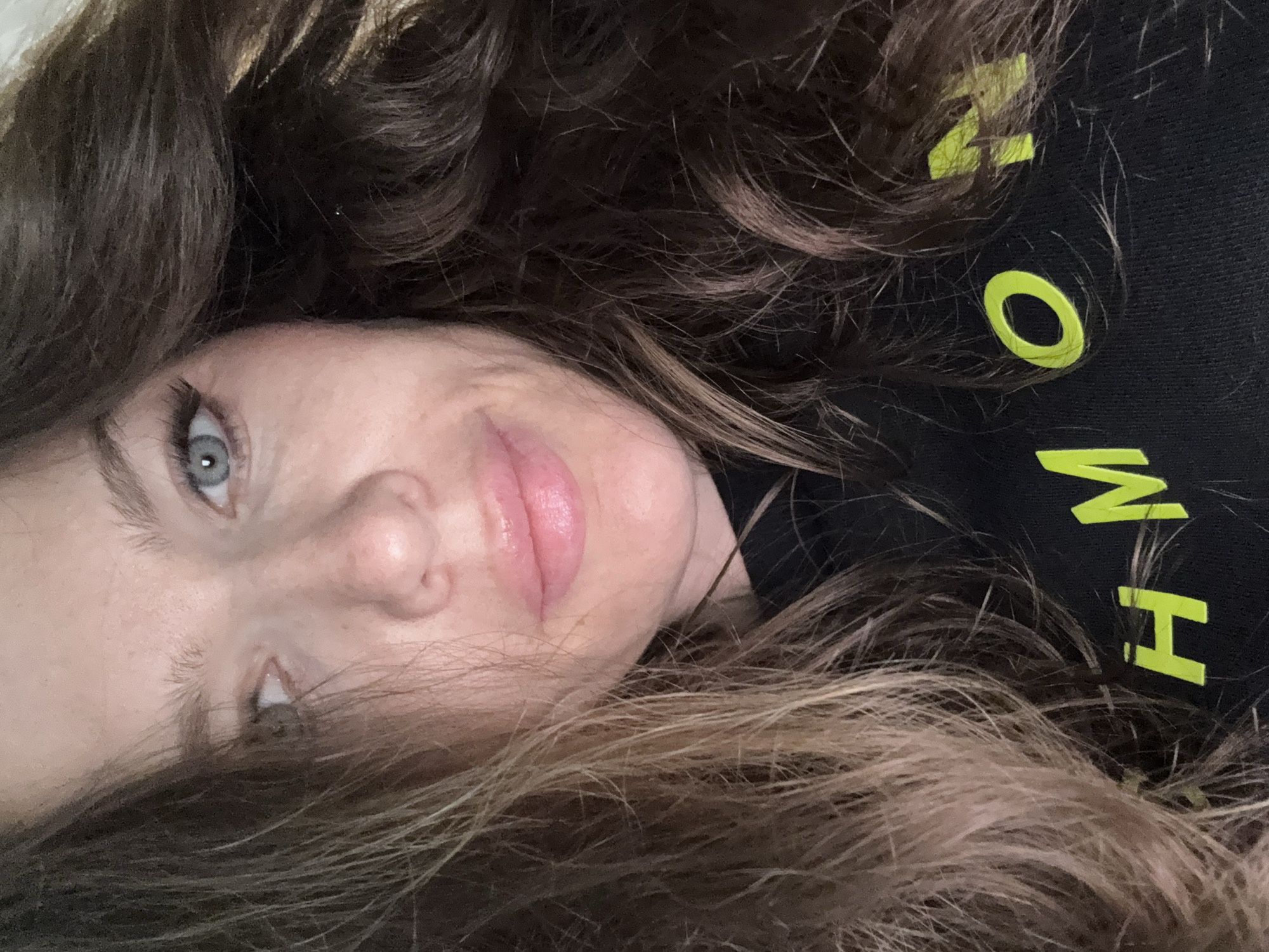 A selfie of a woman with light blue eyes and long, wavy brown and blonde hair. She is wearing a black shirt with yellow letters on it. The letters visible are "MH" and "O". The image is rotated, so she is lying on her side. The location is not identifiable as Moscow, Russia based on the provided image.