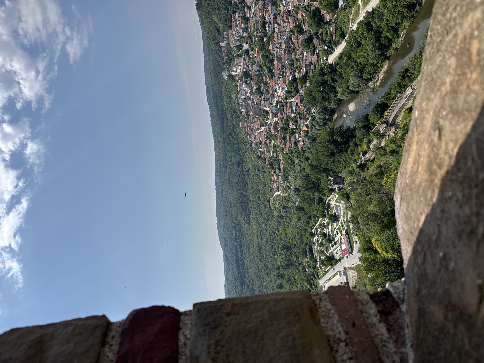 A high-angle, eye-level, wide shot captures a view of Veliko Tarnovo, Bulgaria, from a stone structure. In the foreground, a rough-textured stone wall, with some visible reddish bricks, is out of focus. The midground showcases a dense, green forest covering rolling hills. A winding river flows through the valley, bordered by trees and a section of an ancient stone wall with crenellations. On the opposite side of the river, a town with red-tiled roofs cascades down a hillside, interspersed with white buildings. A road with several cars is visible on the left, adjacent to a park-like area with geometrically arranged gardens. The sky above is a clear blue, with a few scattered white clouds. A small, dark object, possibly an airplane or drone, is visible in the sky.