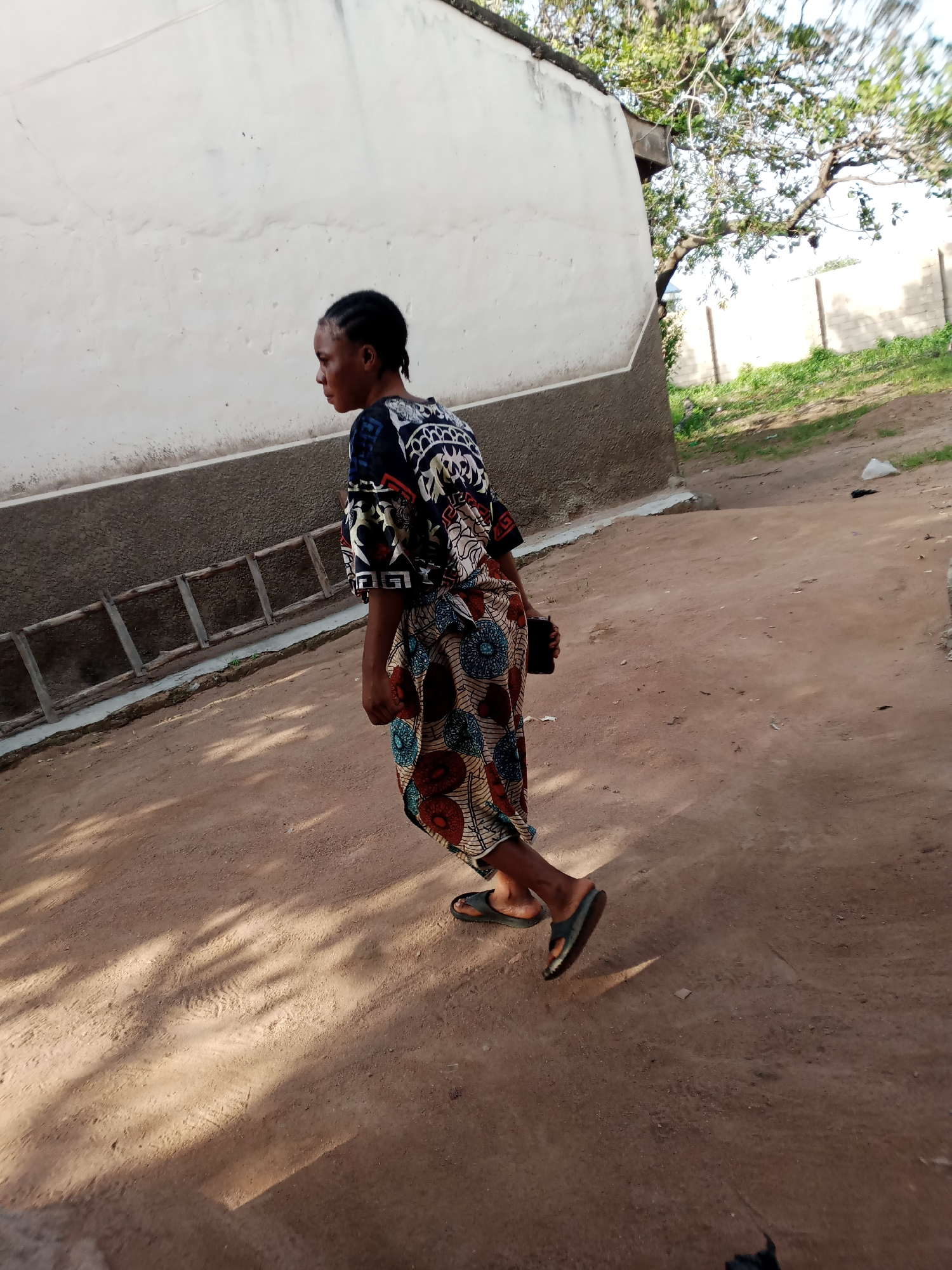 In Jauro Yinu, Nigeria, a young woman walks along a dirt path beside a textured white and brown wall. She wears a vibrant, patterned dress and black flip-flops. Her hair is styled in braids. She is holding a dark object, possibly a phone, in her right hand. A wooden ladder leans against the wall behind her. The ground is a reddish-brown dirt, with shadows cast by sunlight filtering through unseen trees. In the background, a tree with visible branches and leaves extends over a fence constructed of concrete blocks. Patches of green grass are visible near the fence.