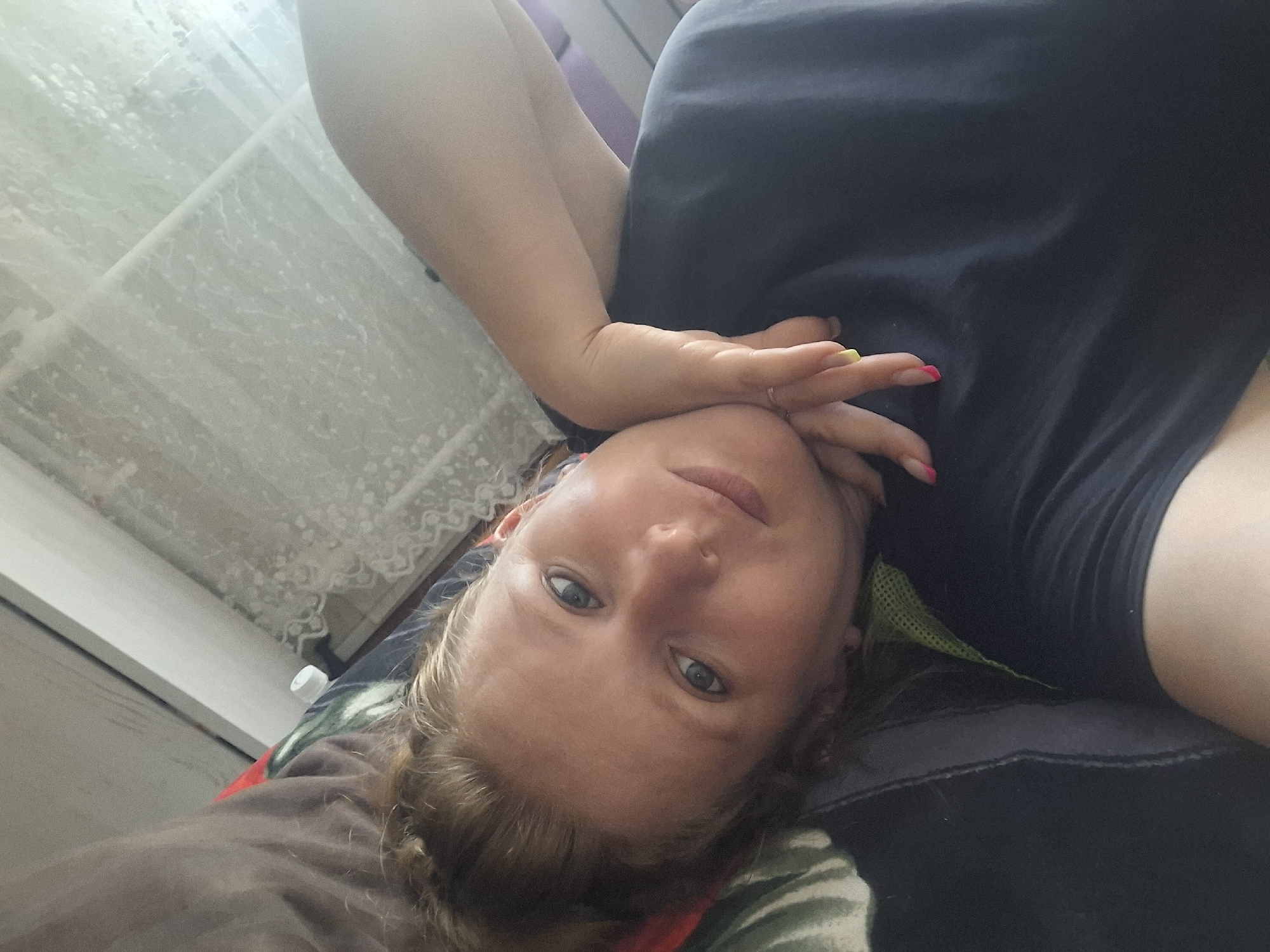 A woman is lying on her back, looking directly at the camera. She has light brown hair styled in a braid, and her eyes are blue. Her left arm is bent, and her hand is resting on her forehead. She is wearing a dark t-shirt and has bright pink and yellow nail polish on her fingers. A thin ring is visible on her right ring finger. The background shows a white patterned curtain and a window. The lighting suggests it is daytime. The image is angled, with the woman's head appearing at the bottom. She appears relaxed and possibly at home.