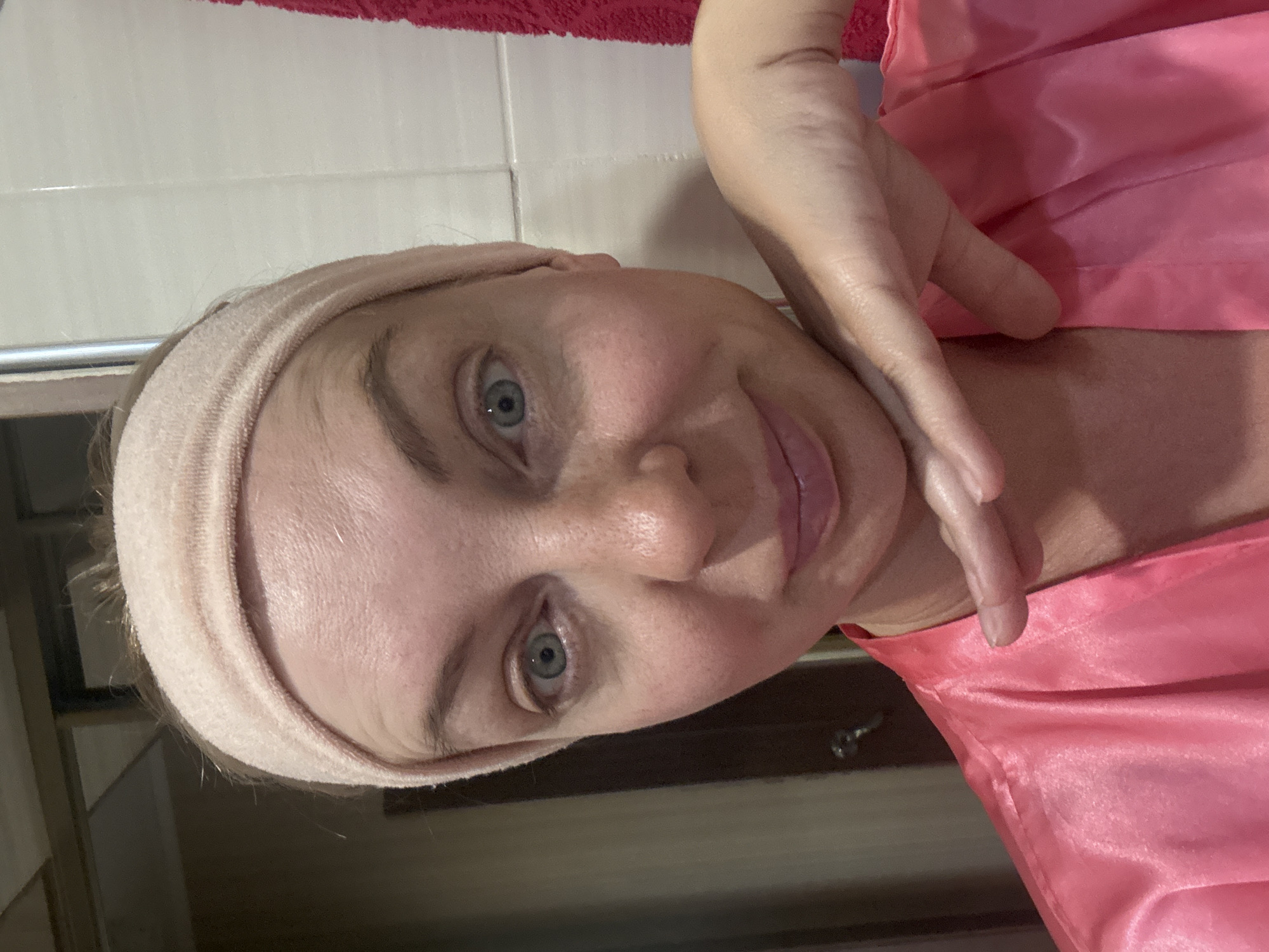 A woman with light skin and blue eyes is looking at the camera. She is wearing a pink satin robe and a light pink headband. Her right hand is resting on her chin. The background appears to be a bathroom with tiled walls. The image is a selfie, likely taken indoors. The location in Sochi, Russia, is not discernible from the image.