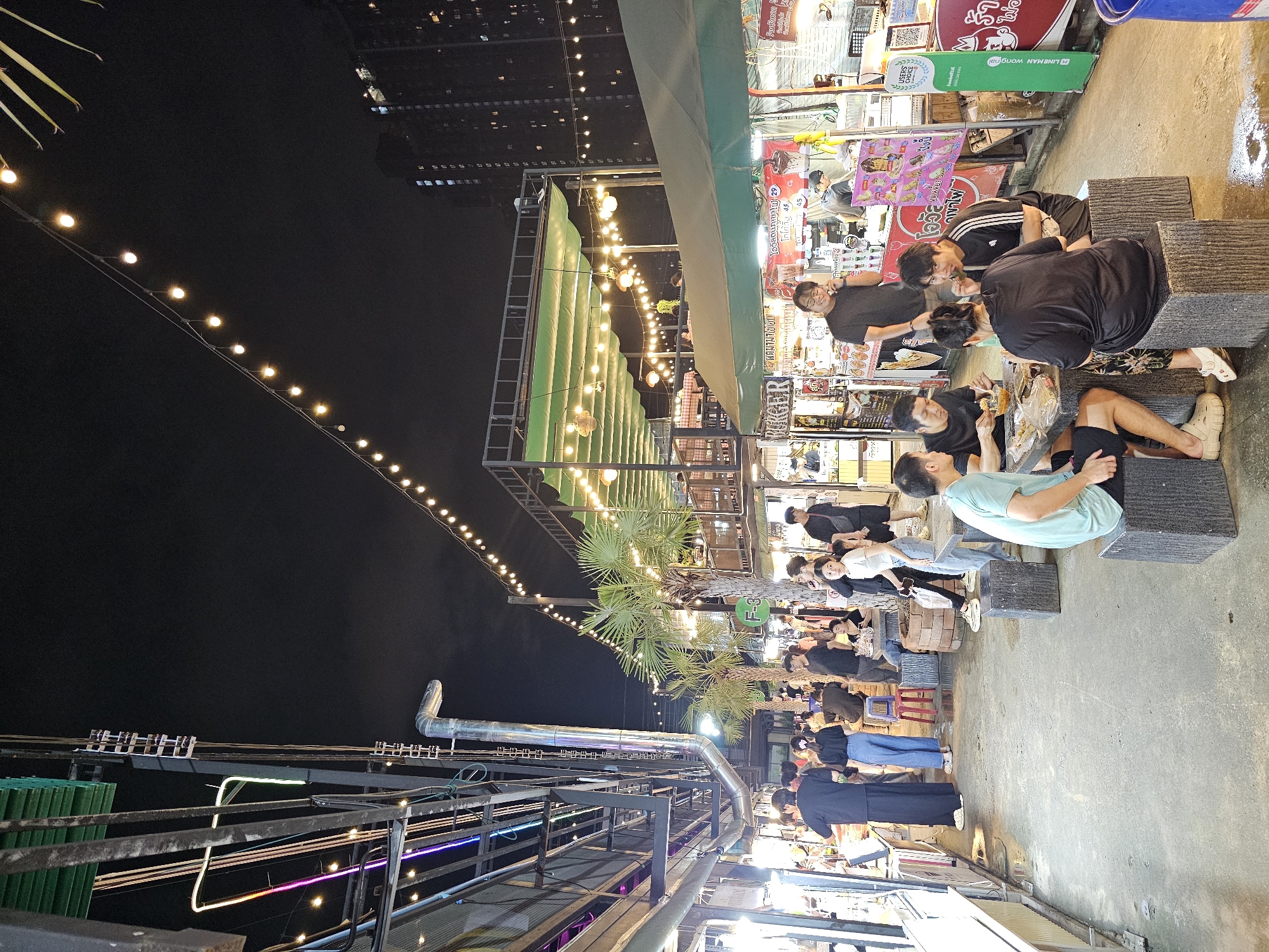 The image depicts a lively night market in Bangkok, Thailand, with a focus on food stalls and outdoor dining. String lights illuminate the scene, casting a warm glow on the bustling atmosphere. Numerous people are seen dining at tables and browsing the stalls, suggesting a popular gathering spot for both locals and tourists. The stalls display a variety of food items, with signage in Thai indicating the types of cuisine offered. The architecture of the market appears to be a mix of temporary structures and more permanent buildings, with some stalls featuring covered seating areas. The presence of palm trees adds a tropical feel to the environment. The overall impression is one of a vibrant and energetic night market experience.