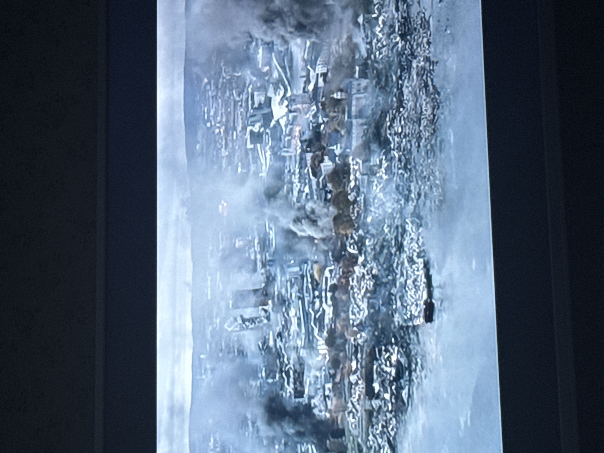 The image shows an aerial view of a city undergoing devastation, likely from an attack or natural disaster. The scene is filled with smoke, obscuring many details. Buildings appear damaged or destroyed, with structural elements visible amidst the debris. The overall environment is bleak and chaotic. The location is not possible to determine based on this image.
