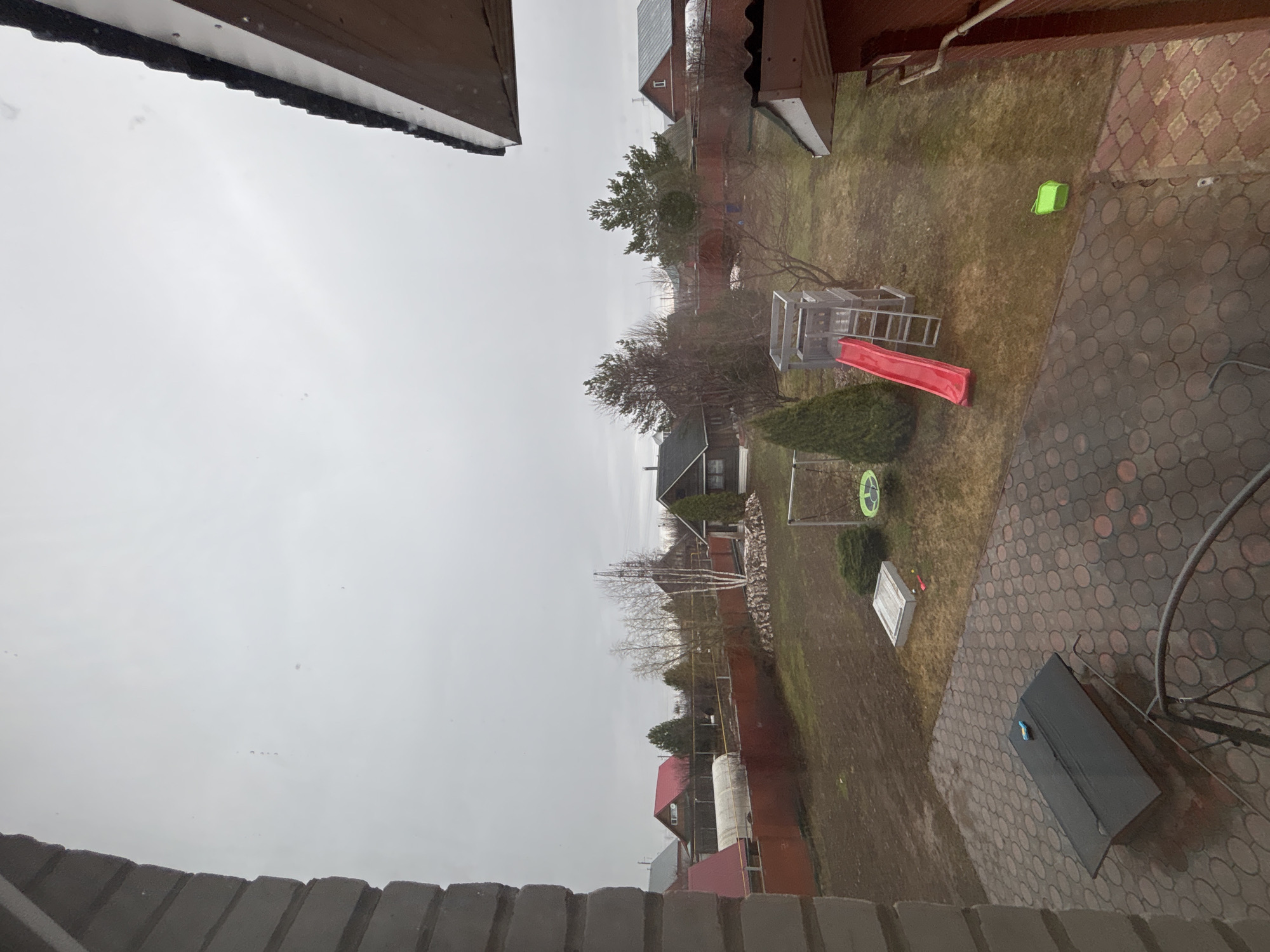 The photograph shows a backyard in Ферма, Russia, under a grey, overcast sky. A children's playground with a red slide is visible. A green bucket sits on the lawn near a brick-paved area, where a dark object is also present. Various houses and trees can be seen in the background, separated by fences. The photograph appears to be taken from an elevated position, possibly from a window. Rain or snow appears to be falling.