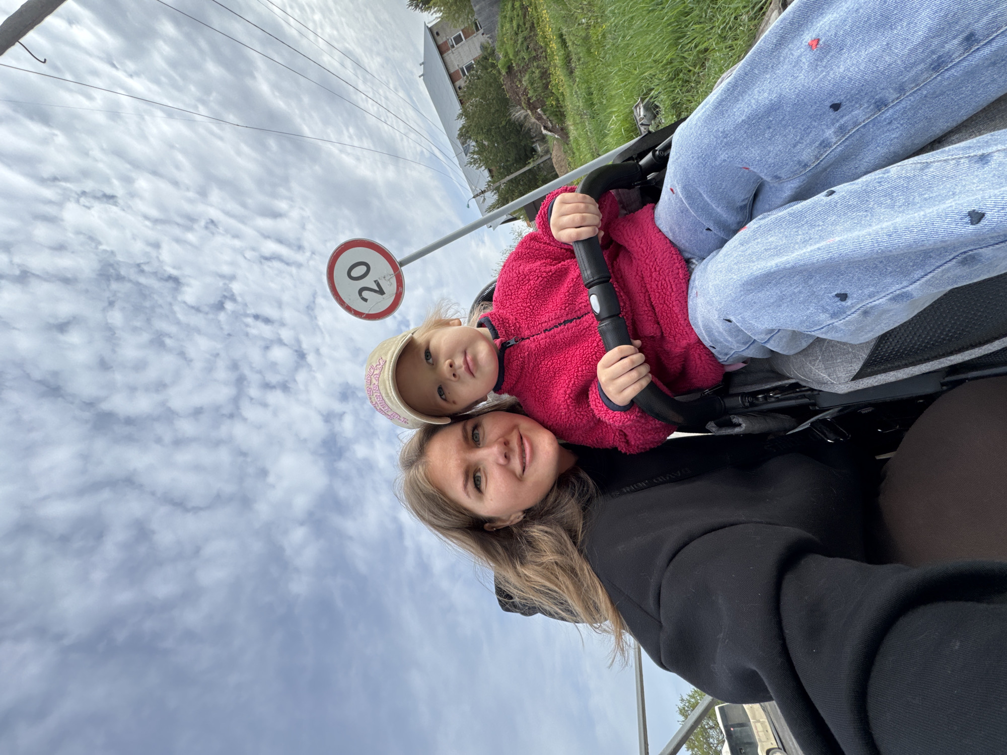 A woman and a young child are positioned in what appears to be a stroller or carriage, captured from a close-up angle. The woman, with light brown hair, wears a black hoodie and faces the camera. The child, who is slightly above her, has a pink jacket and a cap. They are both holding onto the carriage’s handlebar. In the background, a road sign indicates a speed limit of 20. Beyond the sign, there are power lines against a cloudy sky. In the further background, there are green areas and a building. The picture location is in the city of Ферма, Russia.