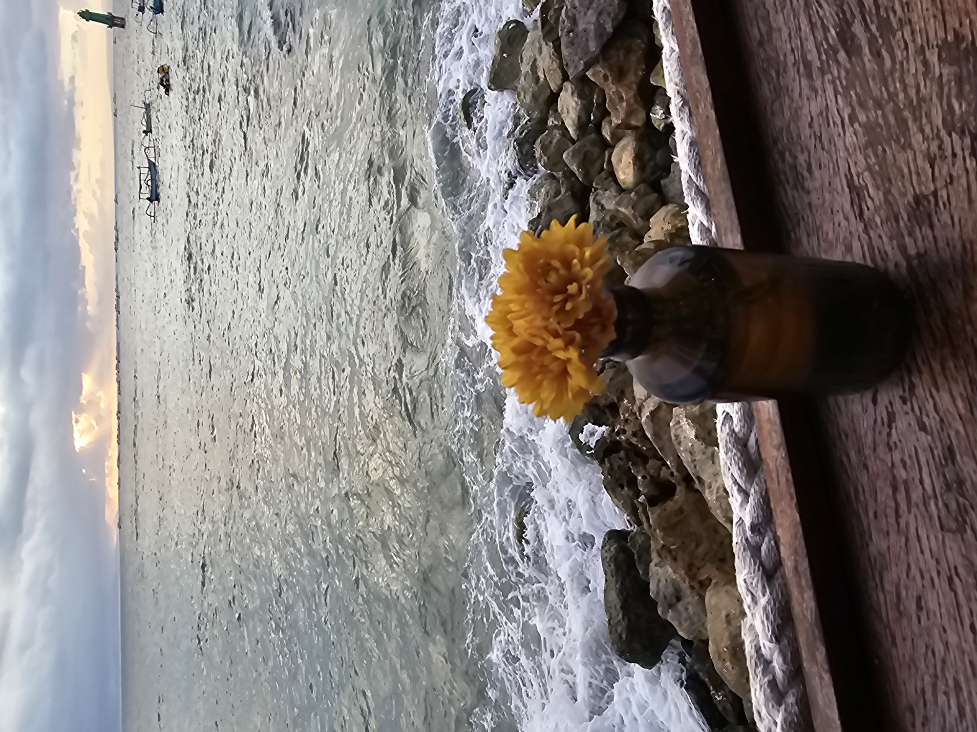The image shows a coastal scene in Denpasar, Indonesia. The focus is on a small vase containing yellow flowers, positioned on a wooden structure overlooking the ocean. The sea is visible, with waves breaking on a rocky shoreline. In the distance, there are several boats, and the sky is overcast with a hint of sunrise or sunset. The overall setting suggests a waterfront area, possibly a restaurant or café, where the floral arrangement adds a decorative element to the seaside ambiance.