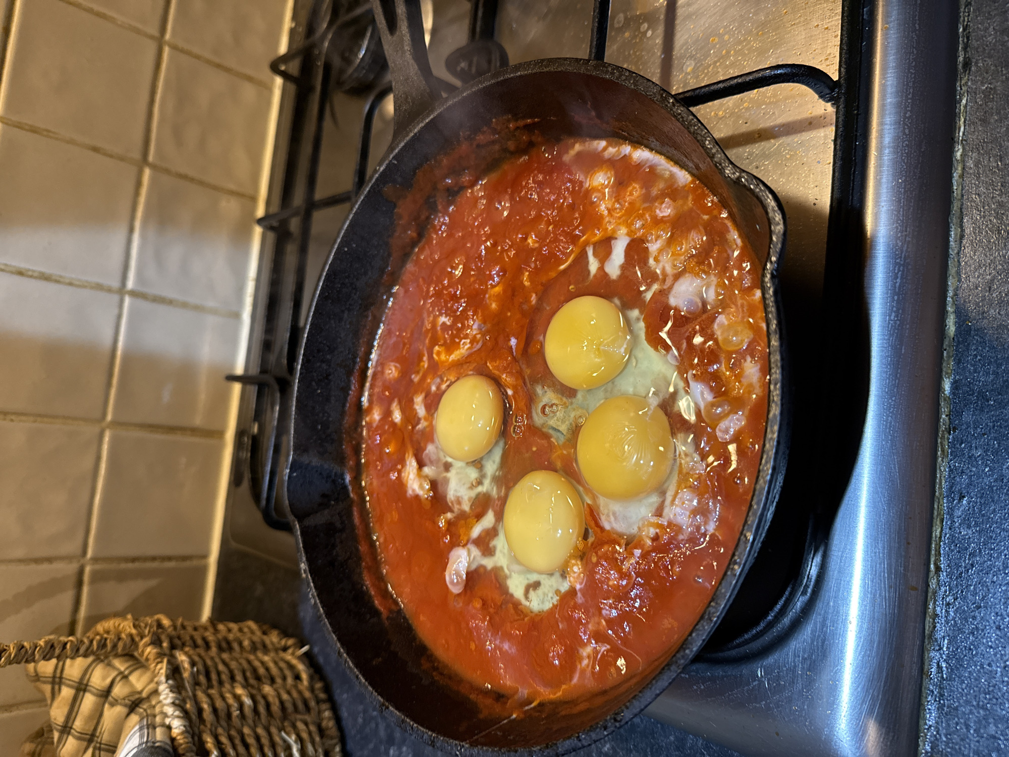 The image shows a cast iron skillet on a gas stovetop, filled with a simmering red tomato sauce with four eggs cracked into it. The stovetop is made of stainless steel. To the left of the stovetop, a tiled wall is visible, as well as a woven basket with a plaid cloth. The image does not contain information to infer location within the city of Great Yarmouth, United Kingdom.