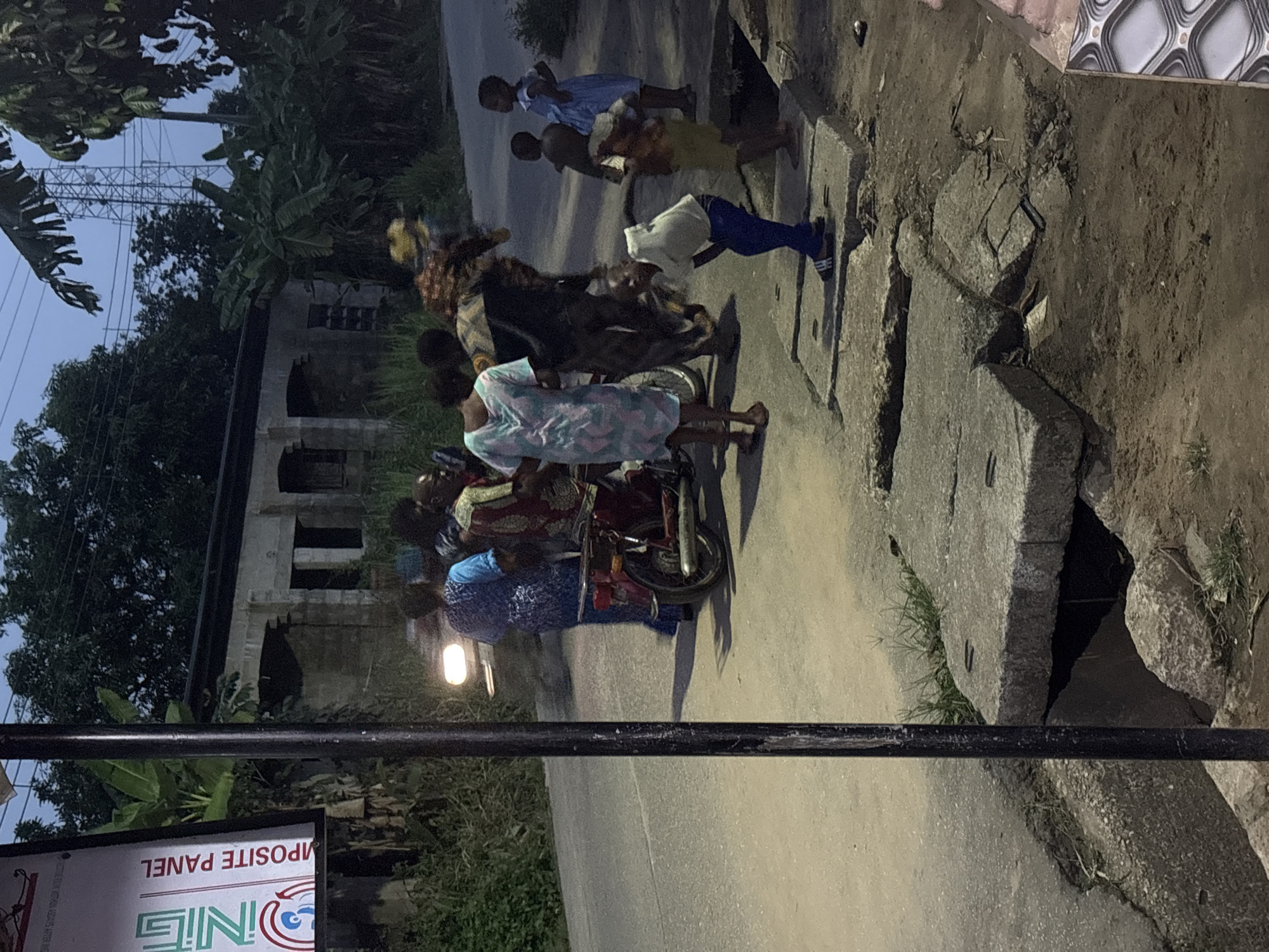 The image shows a group of people standing on a paved road in Ikot Ekpene, Nigeria. Several children, some in school uniforms, are present. Some of the adults are on or near a motorcycle. A building is visible in the background, along with lush vegetation and a utility tower. A sign is partially visible in the foreground. The road surface appears to be concrete or asphalt, with exposed sections and drainage structures along the edge. The lighting indicates it is either early morning or late evening.