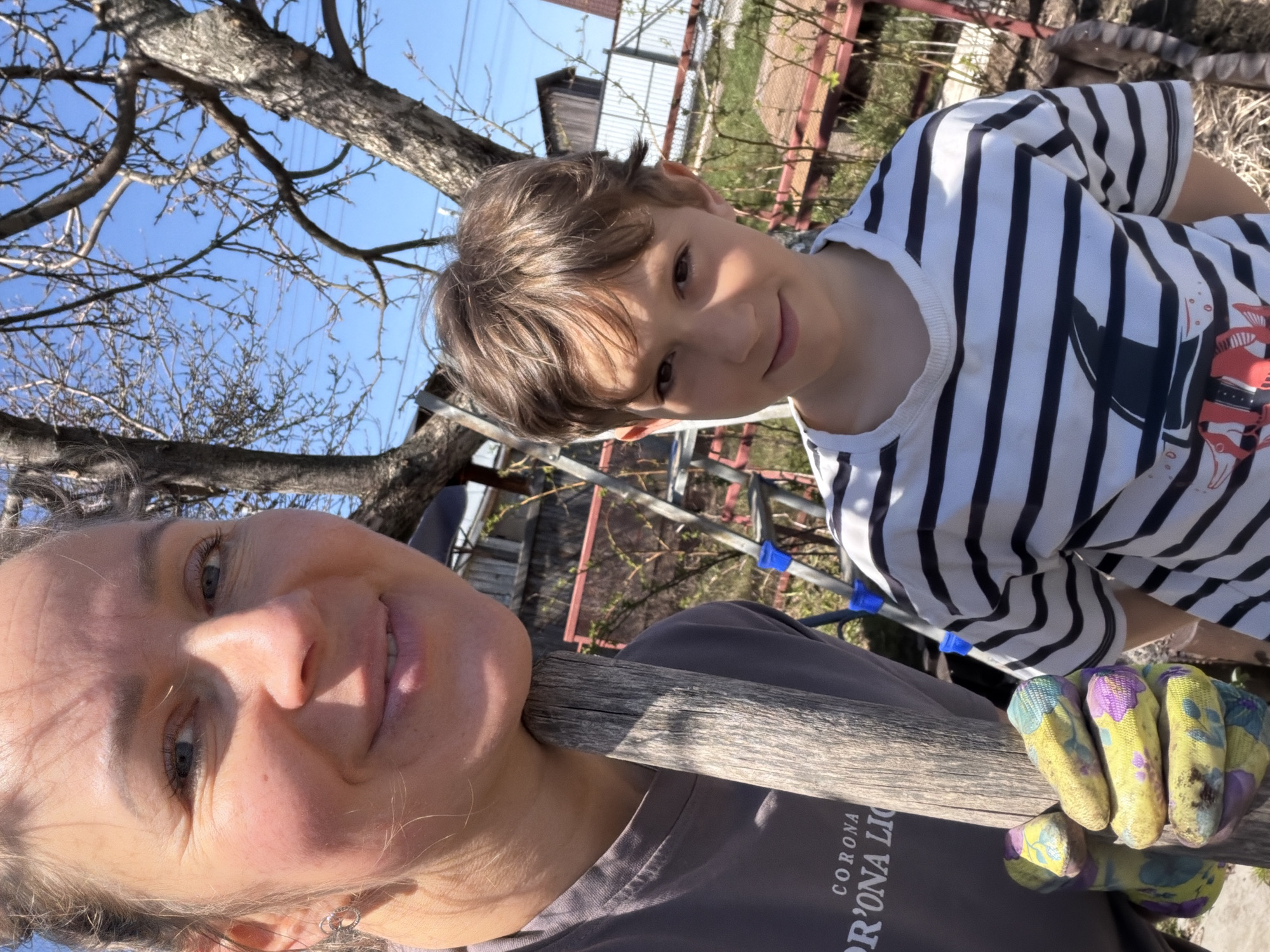 A selfie-style shot shows a woman and a boy outside in what appears to be a garden or yard. The woman is closer to the camera, holding a wooden pole with gloved hands. She has light skin, blue eyes, and a slight smile. The boy stands behind her to the right, wearing a striped shirt and smiling. The background reveals a tree with bare branches against a clear blue sky. In the distance, there's a shed or outbuilding. The presence of gardening gloves suggests outdoor activity, likely related to gardening or yard work. The city of Chaikovsky, Russia, is stated as the location.