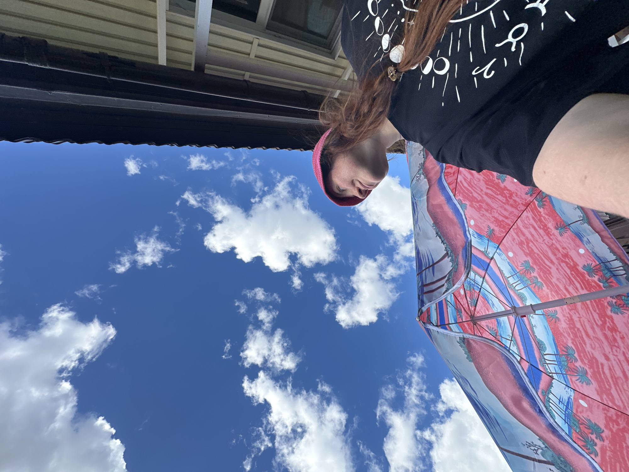 The image, oriented sideways, displays a clear blue sky with scattered white clouds. In the foreground, a person wearing a black t-shirt with white celestial graphics and a pink beanie is partially visible, looking downwards. They are holding up a colorful umbrella with a tropical landscape design featuring palm trees and a pink and blue color scheme. A portion of a building or structure with a light-colored ceiling and dark brown trim is visible at the top of the image, suggesting an outdoor setting, possibly a balcony or patio. There is no discernible information to connect this scene to Nizhny Novgorod, Russia.
