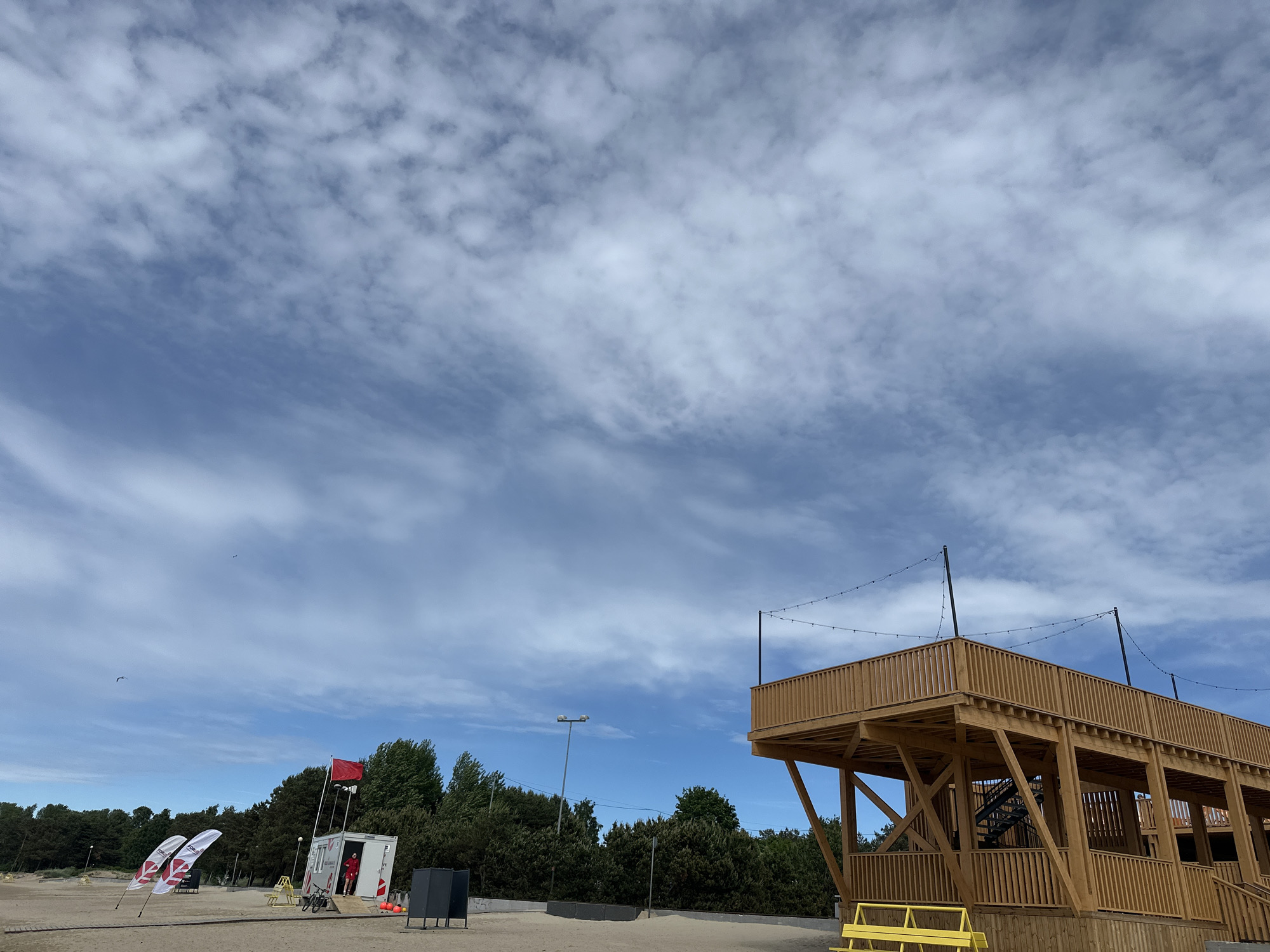 A beachside observation tower made of light-colored wood stands on the right side of the image, its railings and support beams visible against a cloudy blue sky. The tower has multiple levels and an external staircase. In the foreground, a yellow bench is situated at the base of the tower. To the left, a small white building with a red flag on top is present, with two people in red uniforms standing near the entrance. Several white flags with red markings are planted in the sand in front of the building. A line of trees forms a dense backdrop behind the beach structures. The ground is sandy, typical of a beach environment. The overall scene depicts an outdoor recreational area, likely a beach, under a partly cloudy sky.