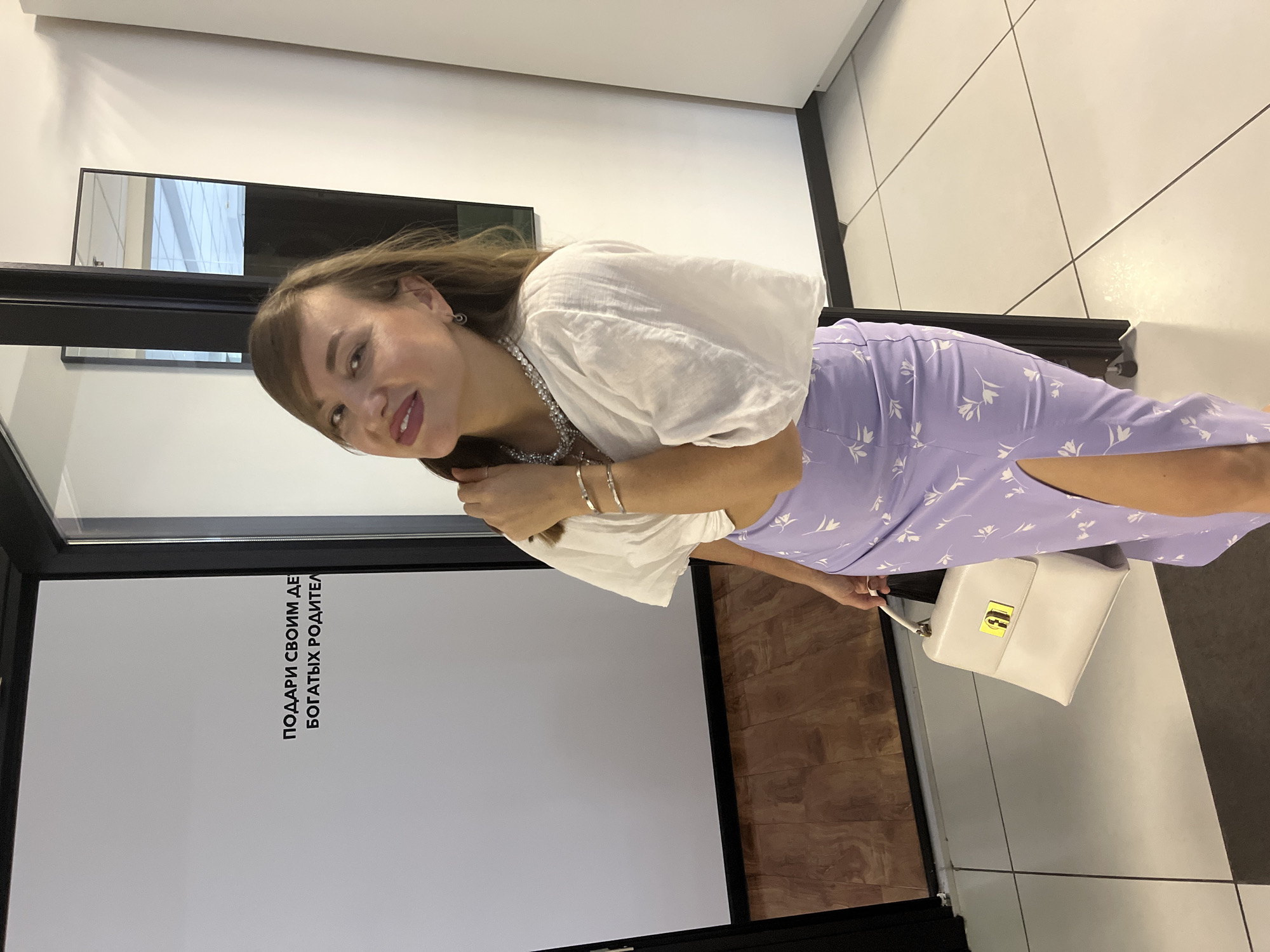 A woman with long brown hair is posing for a photo. She is wearing a white, puffed-sleeved top and a light purple skirt with a floral pattern. She has a necklace and bracelets on. She is holding a white purse with a gold clasp. In the background, there is a white wall with black text and a black frame. There is also a checkered floor and some wooden panels. The woman appears to be smiling and looking at the camera. The text on the wall is in Russian and reads: "ПОДАРИ СВОИМ ДЕТЯМ БОГАТЫХ РОДИТЕЛЕЙ." (Give your children rich parents.) The scene appears to be indoors, possibly in a store or a photo studio. The lighting suggests it is daytime.