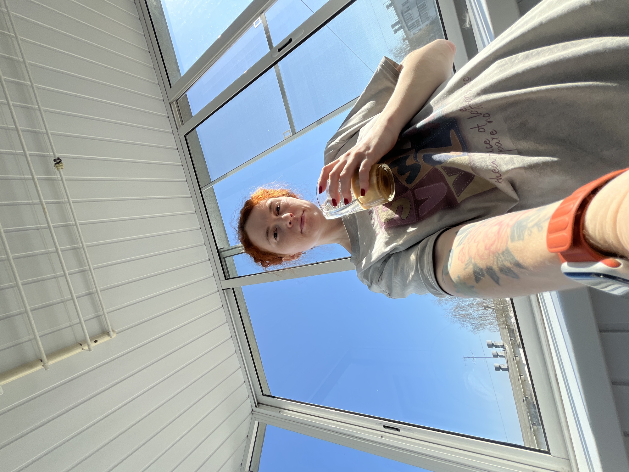 A woman with red hair and a sleeve tattoo holds a glass jar inside a structure with white walls and a glass roof. Outside the structure, a clear blue sky is visible, along with a building and a bare tree branch. The location is the city of Chaikovsky, Russia.