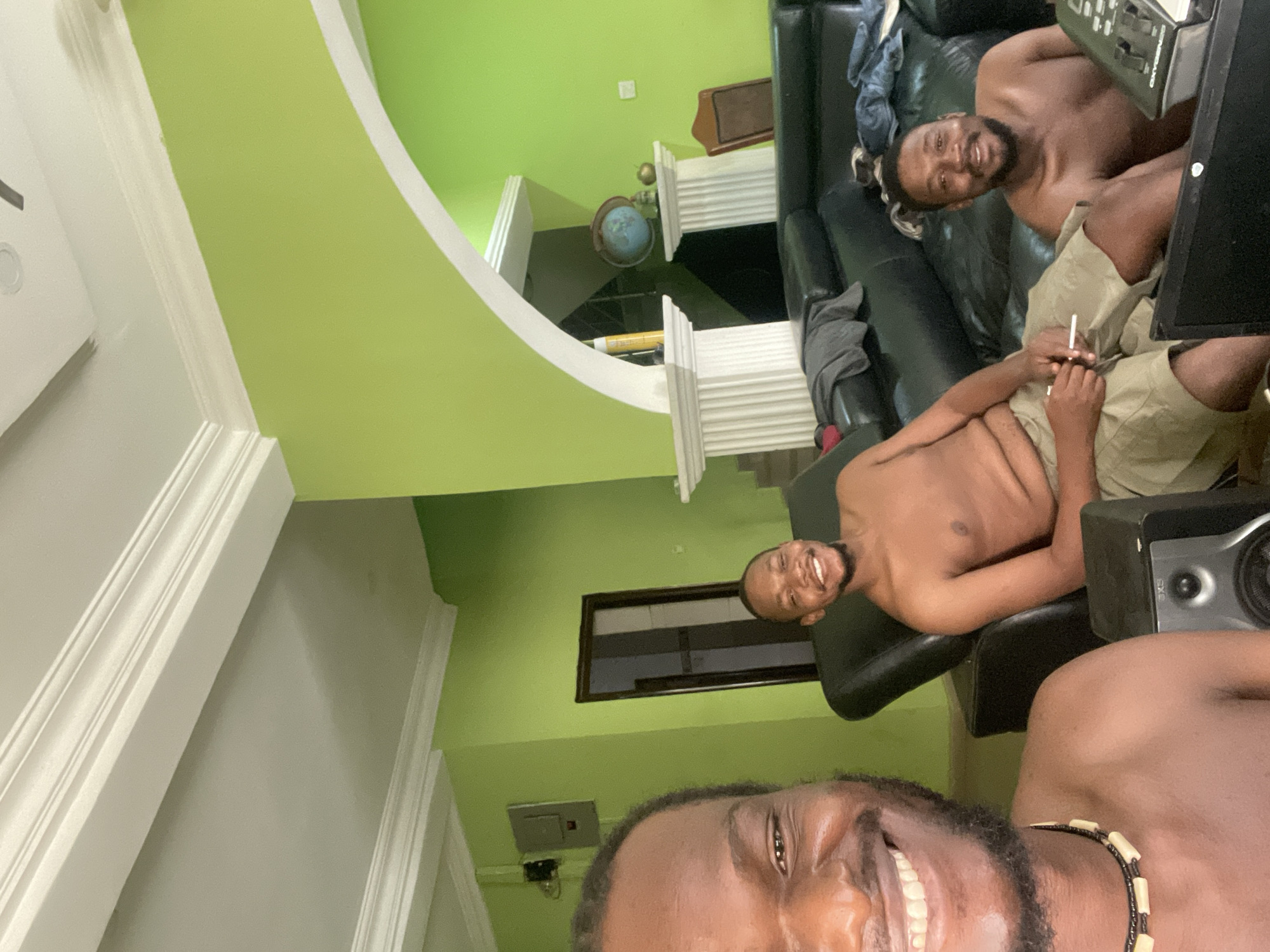 Three shirtless men are in a room with light green walls and white trim. One man is in the foreground, smiling at the camera, wearing a black and white beaded necklace. Behind him to the left, a second man sits on a black leather couch, also smiling. To the right of the second man, a third man reclines on the same couch. The room features a decorative archway with white columns and a globe on display. A dark-framed window is visible in the background. Speakers are in the foreground. The scene is in a residential interior; due to a lack of specific indicators in the image, location details cannot be determined.