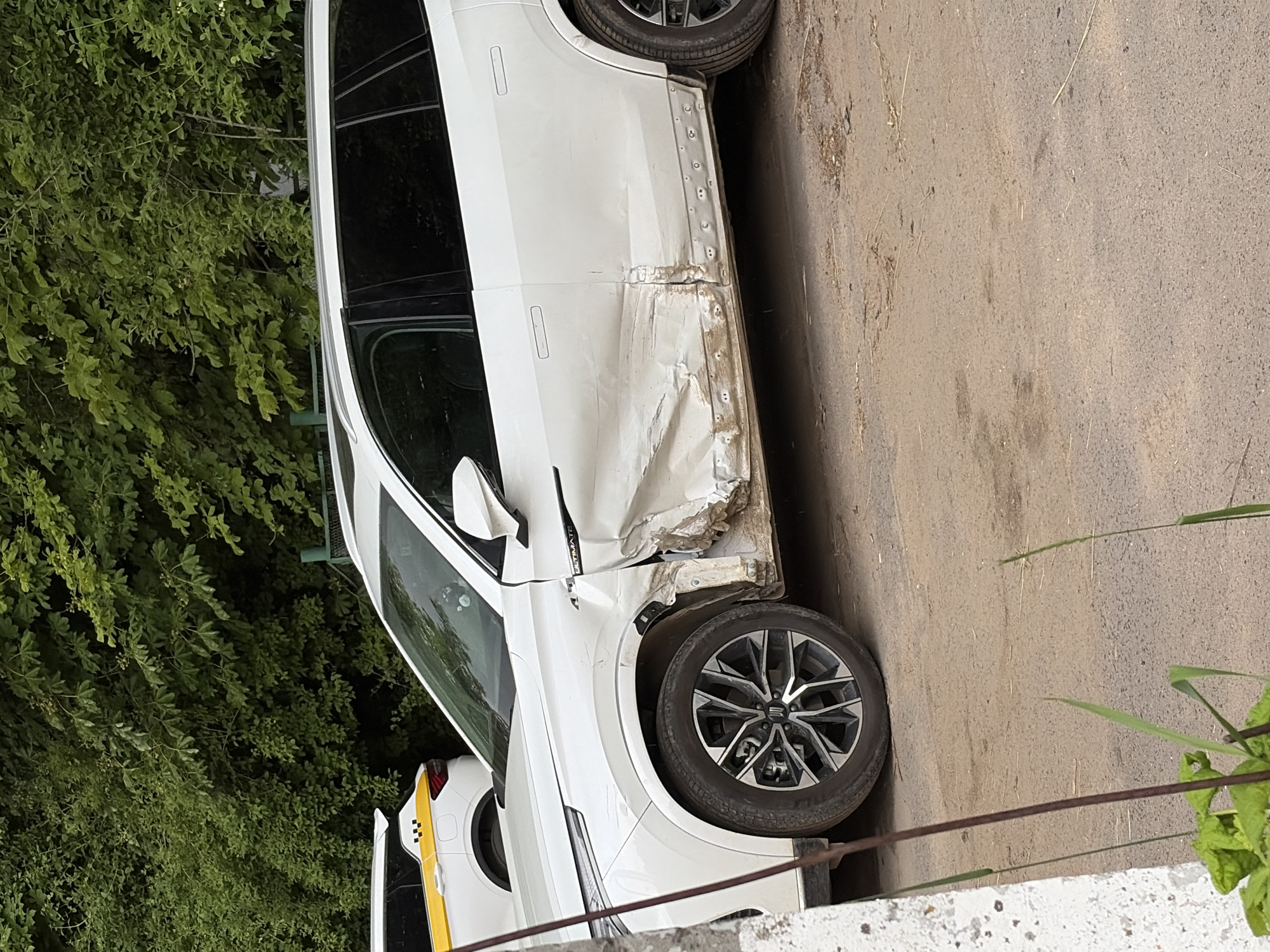 A damaged white SUV is parked on a dirt road in Пикино, Russia. The front passenger side of the vehicle has sustained significant damage, with the door and fender severely dented and creased. The damage extends along the lower body of the car, with visible rivets and rough metalwork on the rocker panel. A yellow and white taxi is parked behind the SUV. Lush green trees form a dense backdrop to the scene. The ground is a mixture of dirt and gravel, with some dry vegetation scattered about. The overall impression is of a vehicle that has been involved in an accident.