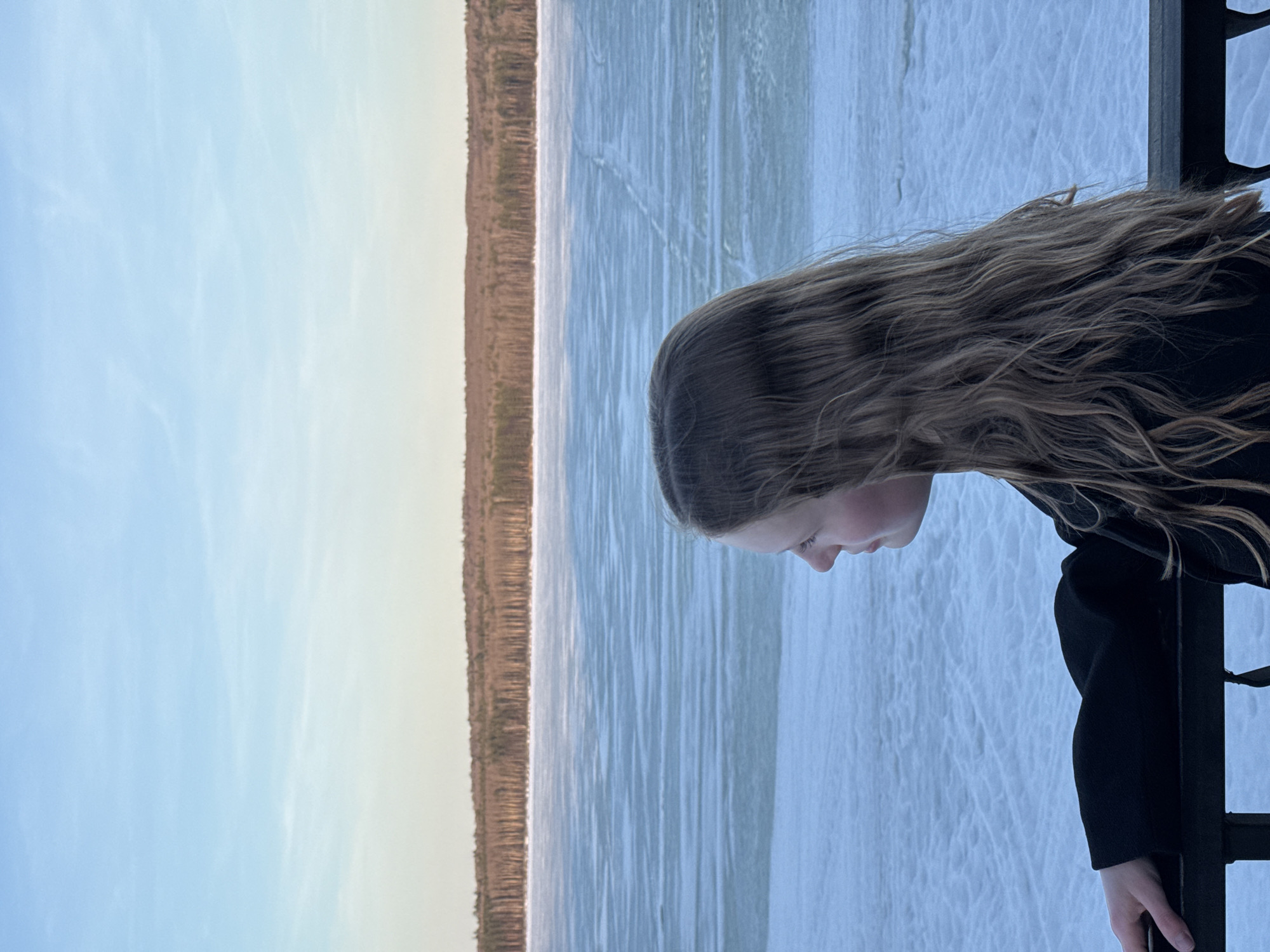 A young girl with long, wavy hair leans over a black metal railing, looking down at a frozen body of water in Chaikovsky, Russia. The water has a light blue tint, and the shoreline across the lake is visible, with a dense line of trees. The sky is a pale blue, suggesting either early morning or late afternoon light. The girl is wearing a dark-colored coat or jacket.