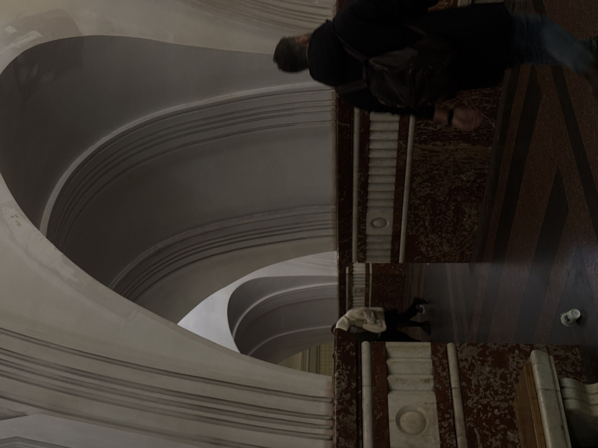The image captures a grand, ornate interior space, likely a public building or a metro station, given the context of Moscow, Russia. The perspective is tilted, making the scene appear rotated clockwise by 90 degrees.
In the foreground, a man is walking across a patterned floor. He is wearing a dark jacket, blue jeans, and a backpack, and his head is visible. The floor features sections of dark brown, possibly marble or patterned carpet, and polished wood. Adjacent to him, there is a detailed architectural structure with reddish-brown and white marble-like elements, including fluted and carved surfaces, indicative of classical or Art Deco design.
Further into the scene, another person or a statue in light-colored clothing is partially visible behind one of these ornate structures. A white disposable cup rests on the floor nearby.
The background is dominated by sweeping, smooth, light-colored architectural curves. These large, graceful forms create a sense of vastness and depth, suggesting vaulted ceilings, archways, or a multi-tiered interior. The design incorporates multiple parallel lines along the curves, adding to the decorative detail. The overall architecture is monumental and elegant, characterized by its smooth, flowing lines and rich materials.
The lighting is diffused, highlighting the architectural details and textures. The scene appears to be indoors, with no direct sunlight visible, suggesting either artificial lighting or indirect natural light. No text is visible in the image.