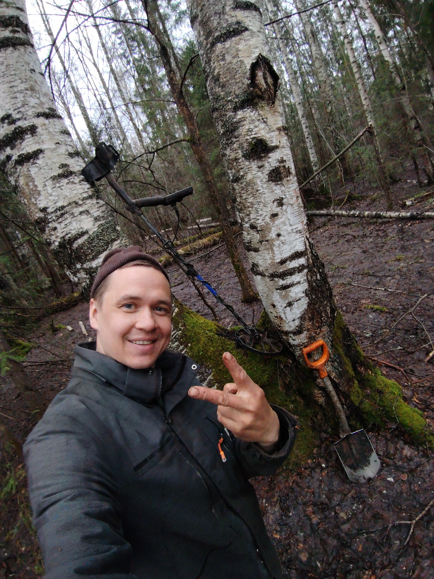 A smiling man takes a selfie in a muddy forest located in Markovskii, Russia. He appears to be in his 30s or 40s, wearing a dark beanie and a dark, practical jacket. He gestures with his right hand, forming a "V" or peace sign.
The setting is a dense forest, characterized by several tall birch trees with white and black bark, along with other bare-branched trees and some conifers visible in the background. The ground is noticeably muddy and covered with fallen leaves and branches, suggesting damp conditions.
To the man's right, leaning against the base of a moss-covered birch tree, are a metal detector and a small shovel with an orange handle. This equipment indicates that the man is engaged in metal detecting. The light is soft and diffused, consistent with an overcast daytime sky. The man's joyful expression conveys a sense of enjoyment and contentment with his outdoor activity. No discernible text is clearly visible on any subjects or objects in the image.