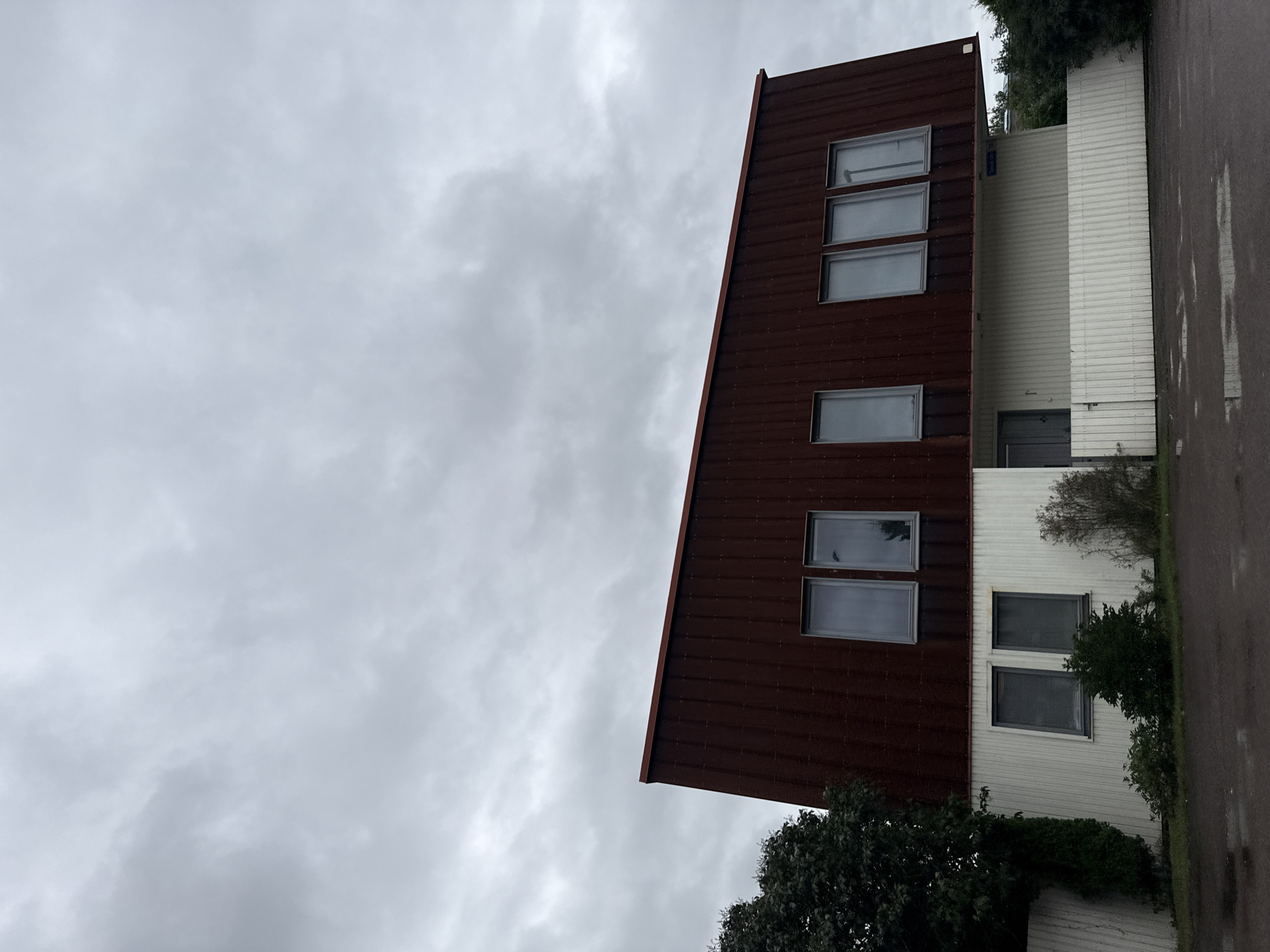 The image shows a two-story building with a distinctive red, sloped roof. The upper section of the building, beneath the roof, is clad in the same red material and features six rectangular windows with gray frames. The lower section of the building is white and appears to be made of vertical panels. It has two windows on the left with gray blinds and a doorway with a gray door on the right, partially obscured by a white fence. There is a white slatted fence running across the front of the lower level. The building is set against a backdrop of a cloudy, overcast sky. To the left of the building, a large, dark green tree is visible. To the right, there are more trees and bushes. The ground in front of the building is asphalt with several puddles, suggesting recent rain. A street sign is partially visible above the white fence on the right side of the building, reading "Suursepä Tänava". This indicates the location is Suursepä Street in Tallinn, Estonia. No people are visible in the image.