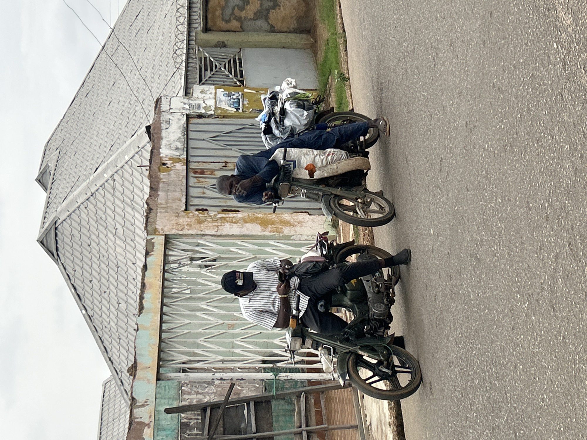 The image shows two men sitting on motorcycles parked on the side of a paved road in Jos, Nigeria. The man closer to the camera wears a striped shirt, dark pants, and a black cap. The other man wears a blue shirt and dark pants. Both motorcycles appear to be older models. Behind the men is a building with a gray roof and metal gates. There is trash and debris piled on the motorcycle farther from the camera.