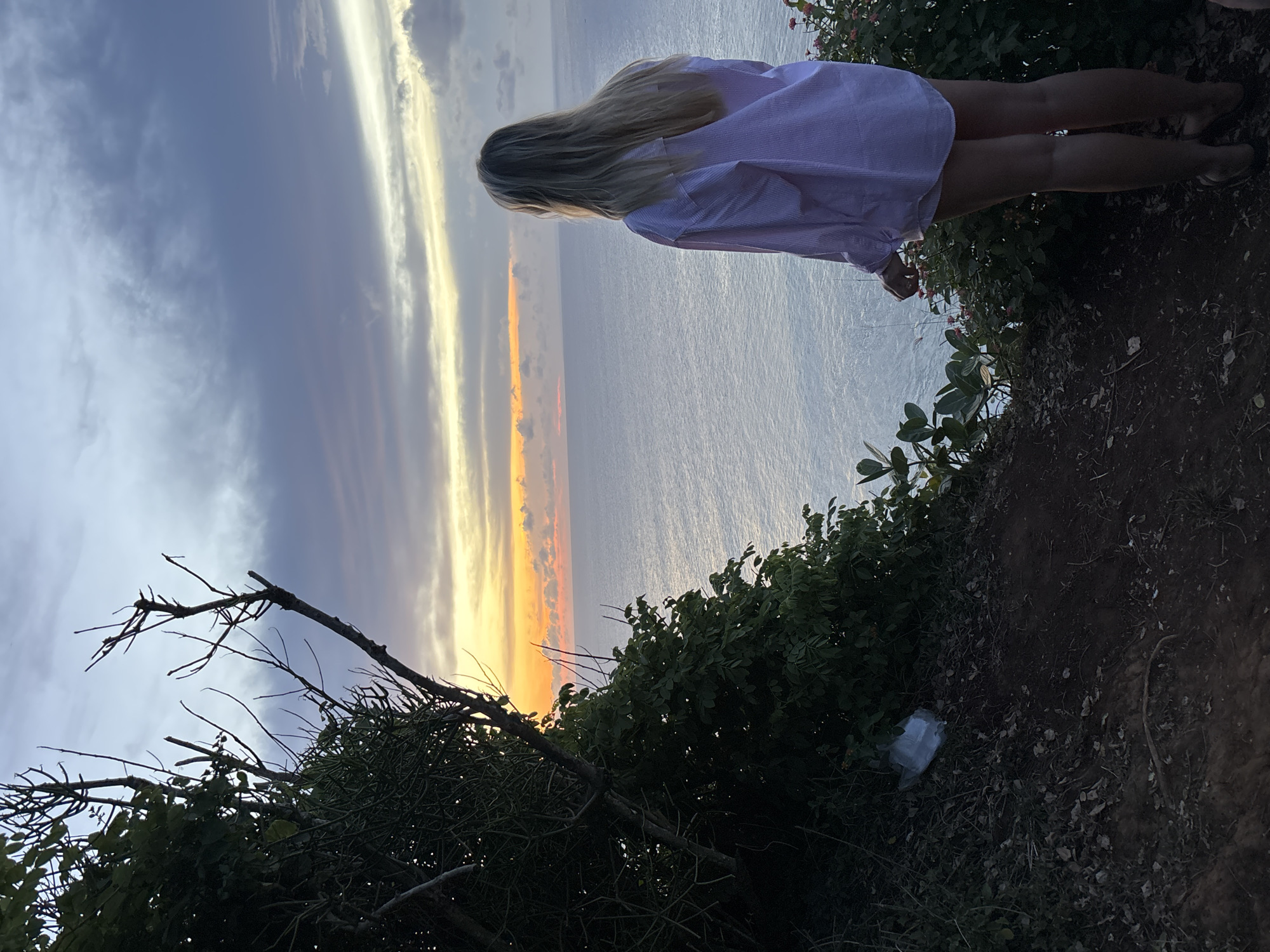 A woman with long blonde hair and a light-colored, button-down shirt stands on the edge of a cliff. She faces towards the ocean, which stretches to the horizon, where a vivid sunset paints the sky with orange and gold hues. The scene unfolds at Uluwatu, Indonesia, characterized by its elevated coastal terrain and lush tropical vegetation. The cliff's edge is covered in greenery, with trees and shrubs framing the view. The ground is primarily soil and leaf litter, with a discarded plastic bag visible.
