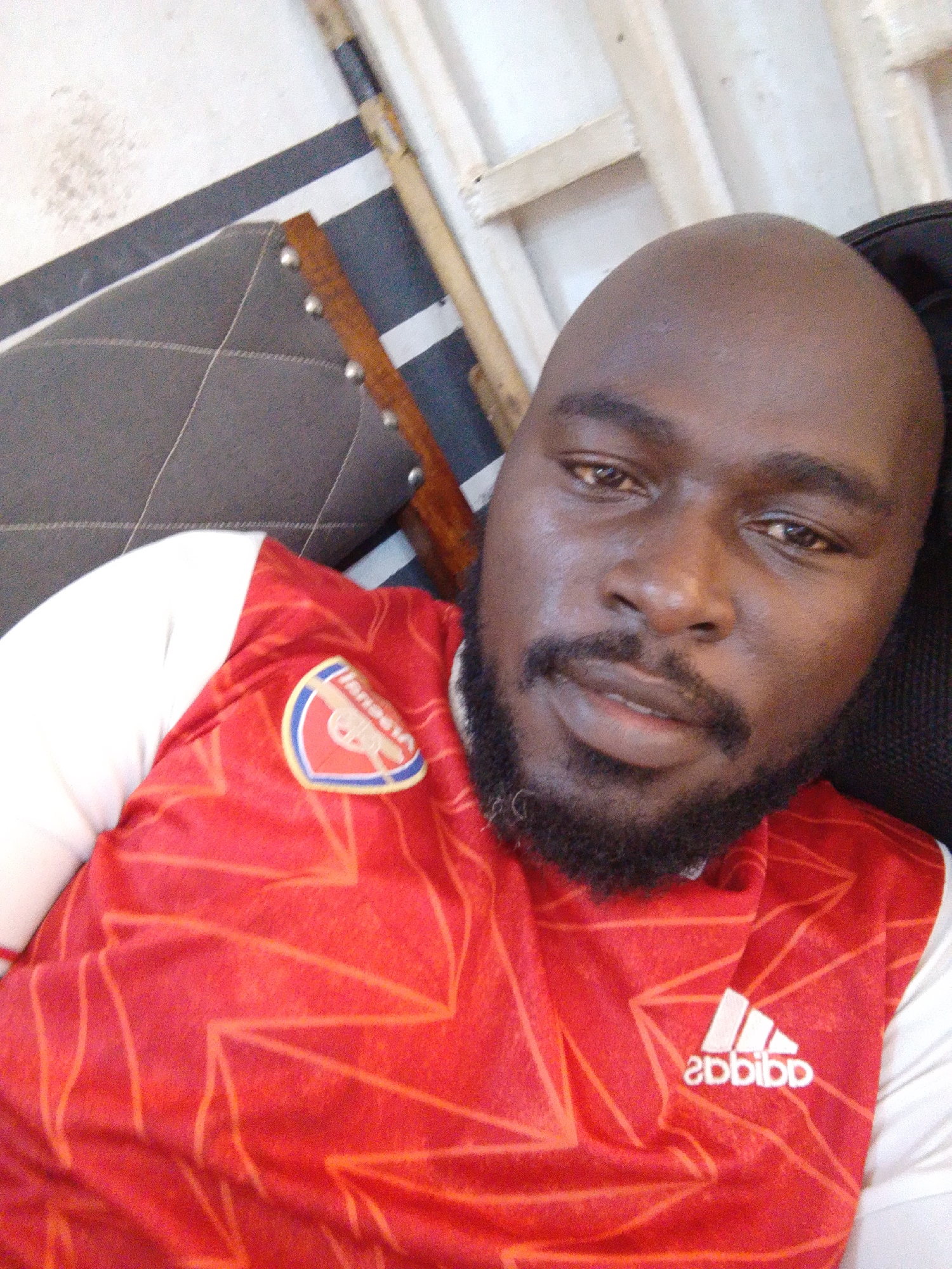 A close-up photograph depicts a dark-skinned man with a bald head and a beard, wearing a red and white Arsenal soccer jersey with an Adidas logo. The background includes a dark gray textured surface, possibly part of a chair or couch, and a wooden structure or object in the upper portion. The location is unknown.