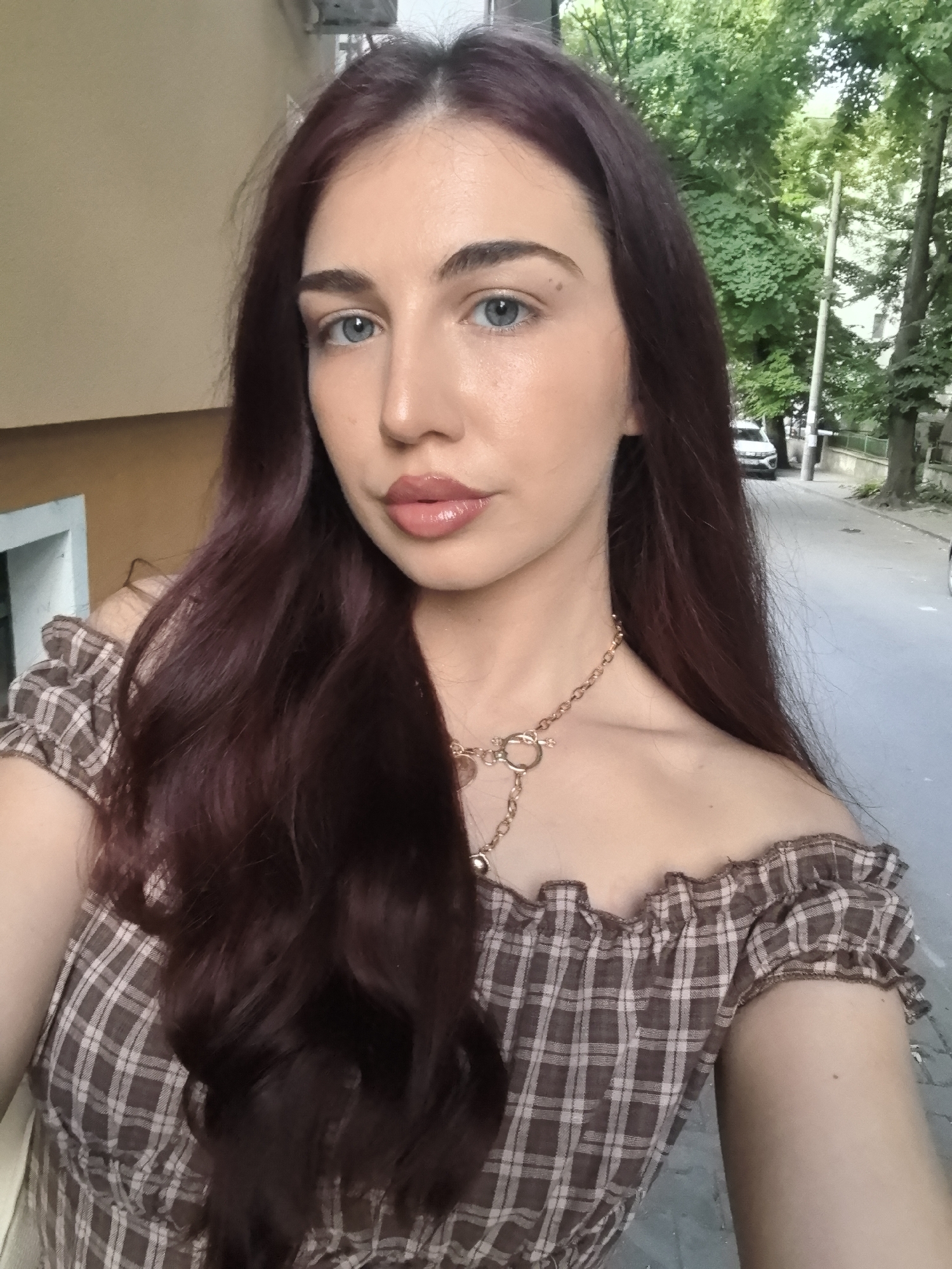 A young woman with long, dark auburn hair and striking blue eyes takes a selfie outdoors. She is wearing an off-the-shoulder, plaid top with ruffled sleeves and a gold chain necklace. The background suggests a street scene in Stara Zagora, Bulgaria, with trees lining a sidewalk and a car visible in the distance. The lighting indicates it is daytime.