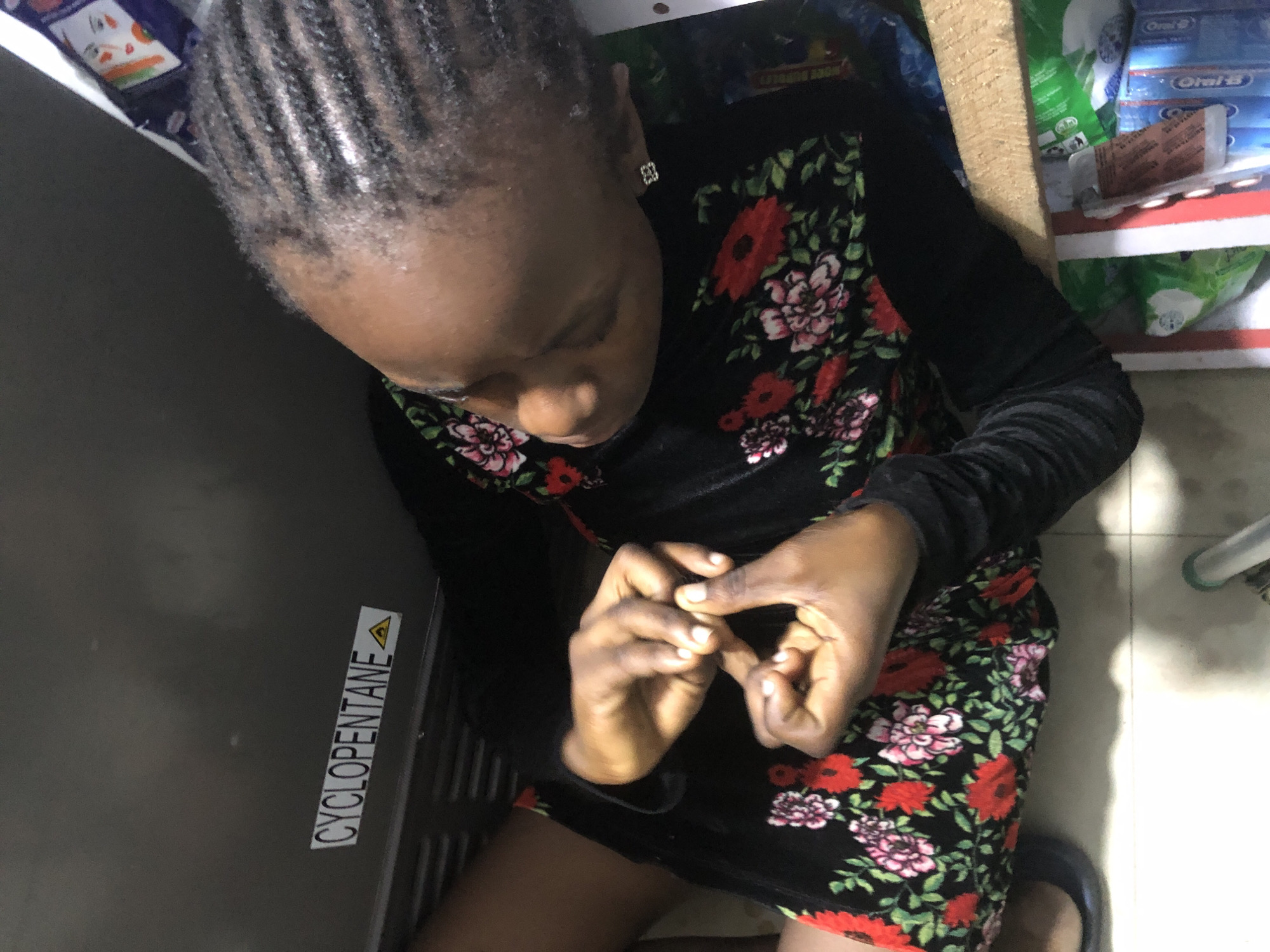 A young Black female is sitting on the floor. She is wearing a black dress with a floral print of red and pink flowers. Her hands are clasped together in her lap. She has braided hair. In the background, there is a metallic surface with a sticker that reads "CYCLOPENTANE". There are also some boxes and packaging on a wooden shelf. The flooring appears to be tiled. The scene is likely an indoor domestic or commercial setting.