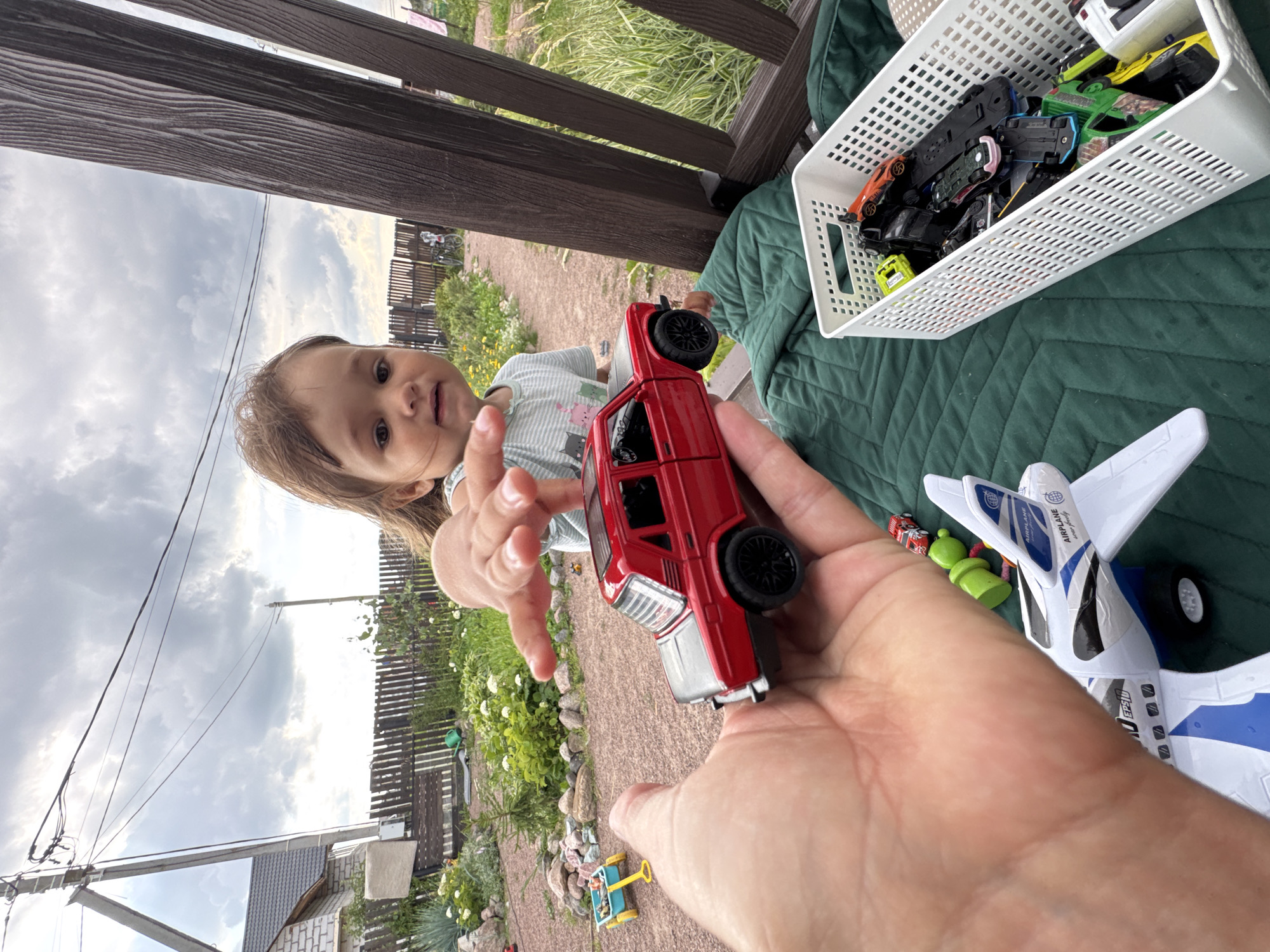 In Kempelevo, Russia, a baby reaches for a red toy car held by an adult's hand in the foreground. The baby, wearing a striped t-shirt, is looking towards the car with an outstretched hand. Behind them, a green quilted surface is visible, with a white plastic basket filled with various toy cars. A large white toy airplane is positioned on the green surface, partially obscuring the adult's hand. The background depicts a rural setting with a wooden fence, lush green vegetation, and a cloudy sky overhead. A house with a light-colored brick exterior and a pitched roof is visible to the left, along with utility poles and wires.