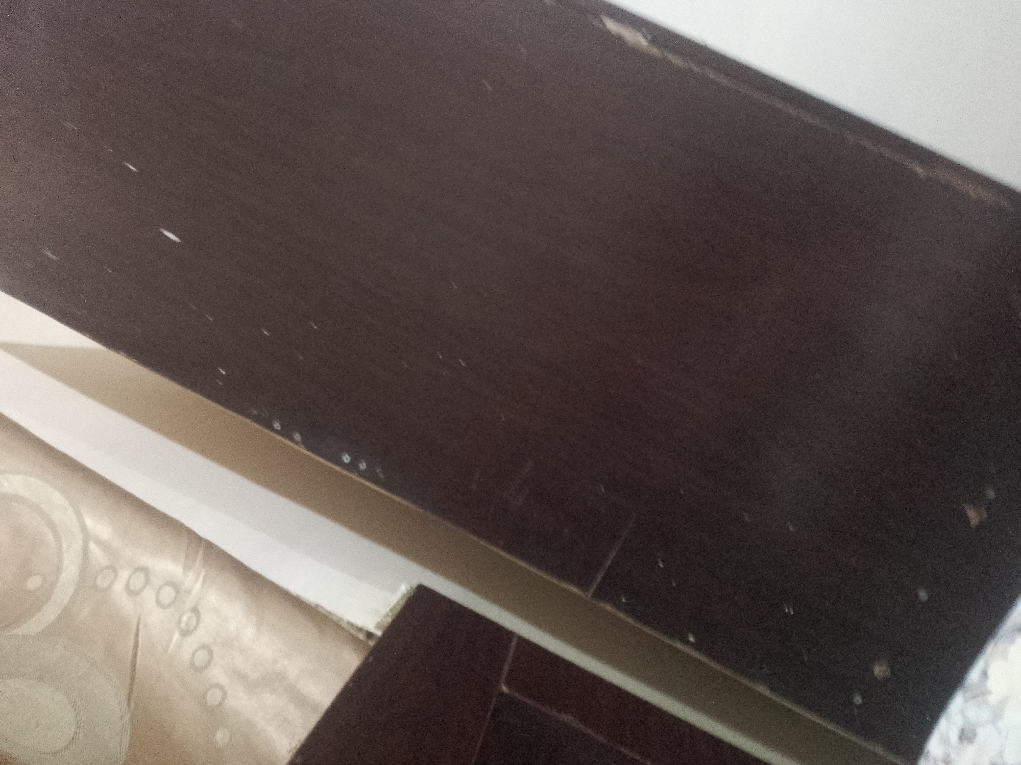 The image shows a close-up view of a dark brown wooden surface, likely a shelf or cabinet, angled from the top left to the bottom right of the frame. The surface has a matte finish and is textured with visible wood grain. There are numerous small white specks scattered across the surface, possibly dust or minor damage. A lighter-colored, possibly fabric material with a circular pattern is visible in the bottom left quadrant of the image, partially obscured by the dark surface. The background is a plain, light-colored wall. The lighting appears to be from the left, casting subtle shadows and highlighting the texture of the wood. The scene is indoors, with no visible signs of the city or specific location.