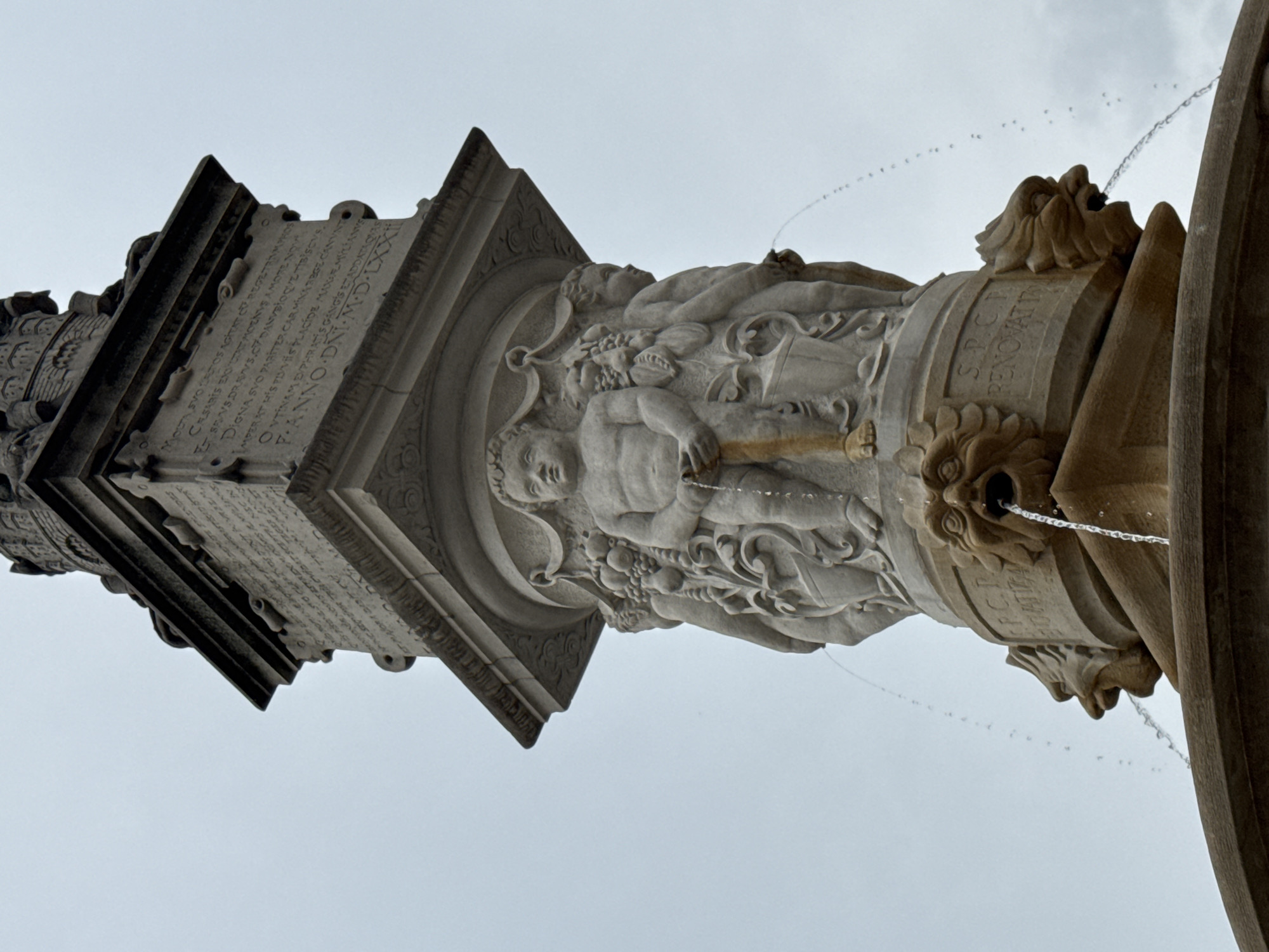 This image shows a close-up, low-angle view of the Roland Fountain (also known as Ganymede's Fountain) located in Bratislava, Slovakia. The fountain is made of stone and features several sculpted elements. In the foreground, two grotesque lion-like heads are visible, with water spouting from their mouths, forming streams that fall into an unseen basin. Above these heads, the central part of the fountain depicts several putti (cherubic figures) and decorative carvings. One central putto, with a muscular physique, holds a wooden object. Water also streams from the hands of other putti located on the sides. The upper section of the fountain features a stone pedestal with carved inscriptions in Latin. The sky is overcast and appears in the background. The image is taken from a street-level perspective, focusing on the details of the fountain.