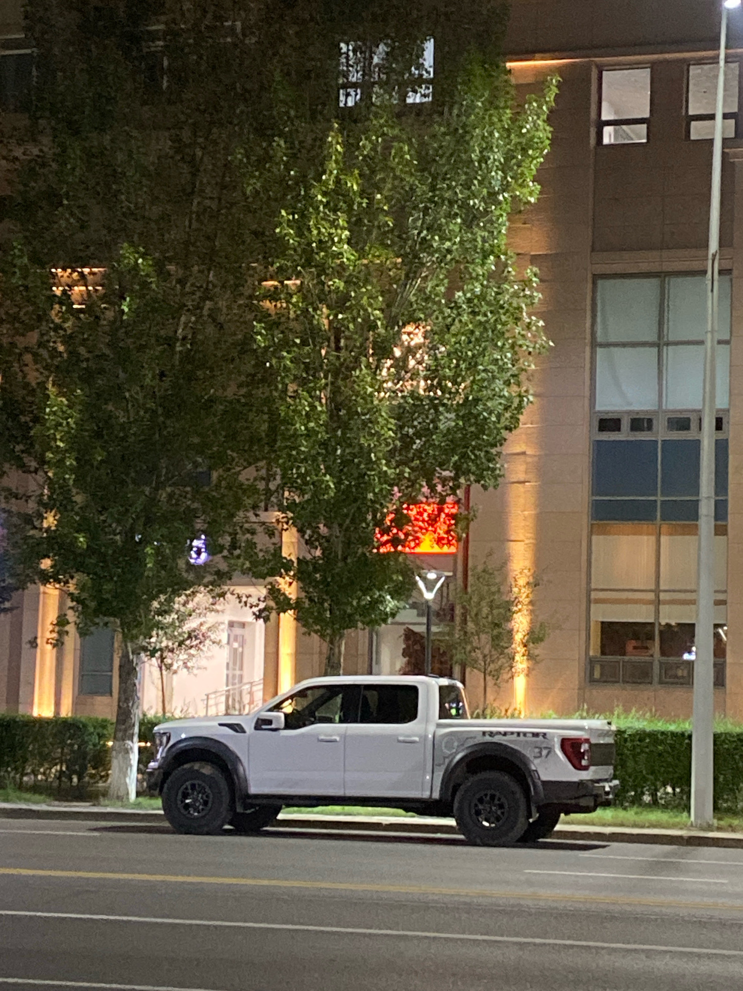 A white Ford F-150 Raptor pickup truck is parked on a street in Astana, Kazakhstan, with a modern building and trees in the background. The truck is facing left, and its rear is partially visible. The building features large windows and is lit from within. The scene is captured at night, with streetlights and building lights illuminating the area. The truck has black wheels and "RAPTOR 37" decals on its side.